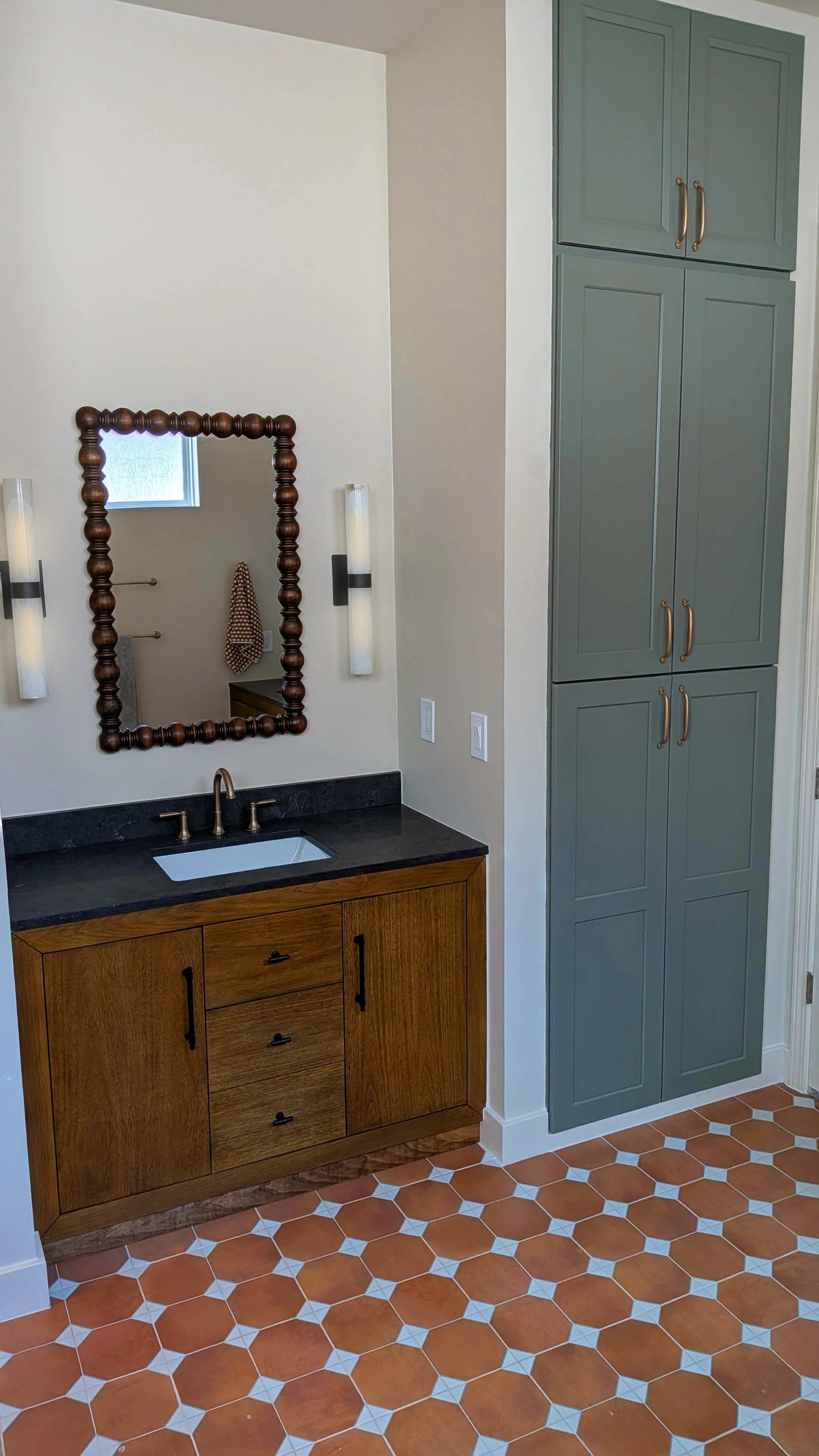 Bathroom vanity with a black countertop, a white undermount sink, and a wooden cabinet with six drawers. A large mirror with a wooden beaded frame hangs on the wall, flanked by two wall-mounted light fixtures. To the right, there are tall, green, paneled cabinets with gold handles. The floor is covered with reddish-brown hexagonal tiles with white accents.