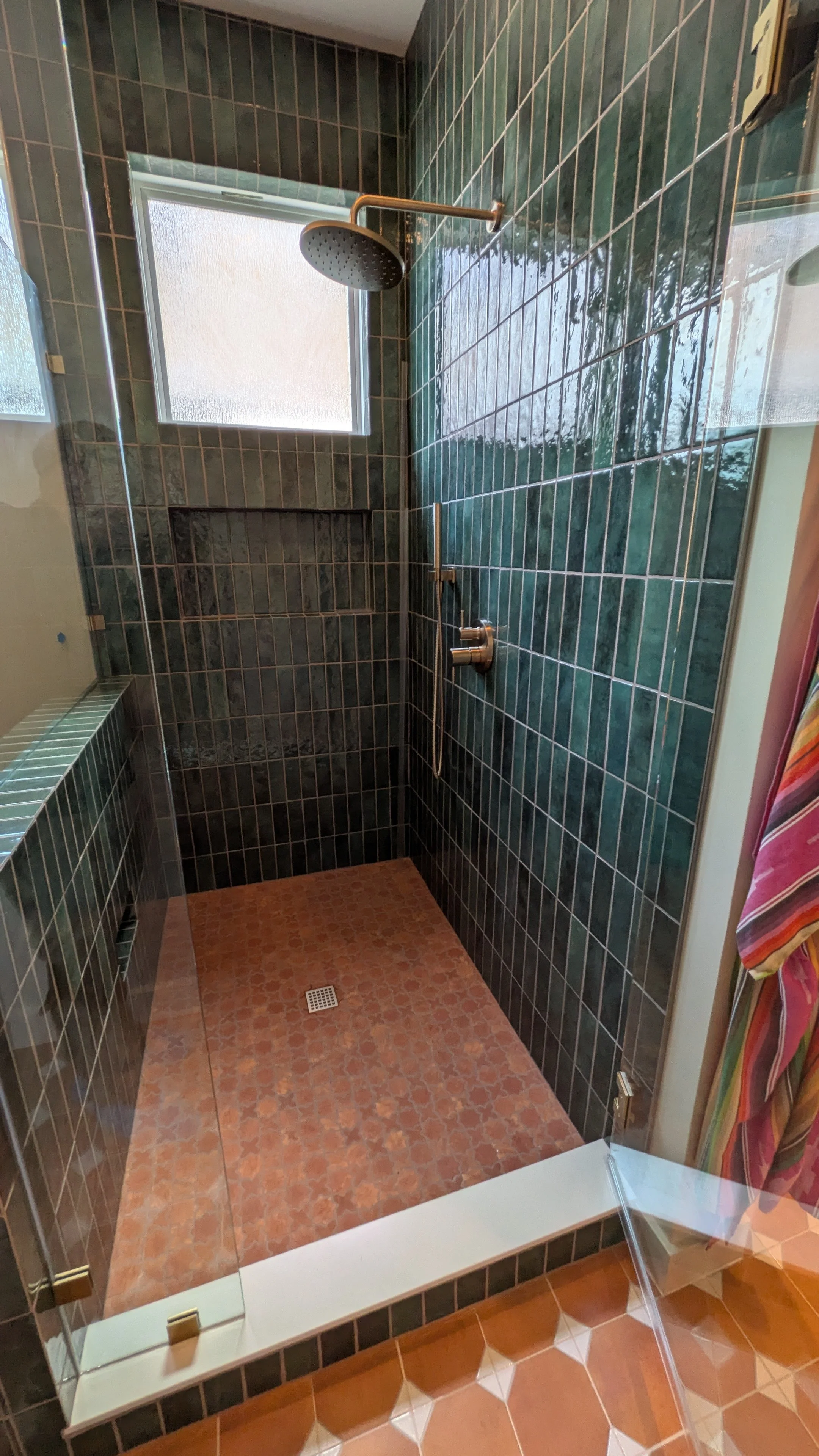 A walk-in shower with dark green tiled walls, a window with frosted glass, a round rain showerhead, and a small built-in shelf. The shower floor has small reddish-brown tiles, a drain, and is enclosed by a glass wall with brass-colored handles. Multicolored striped towel hangs outside the shower.