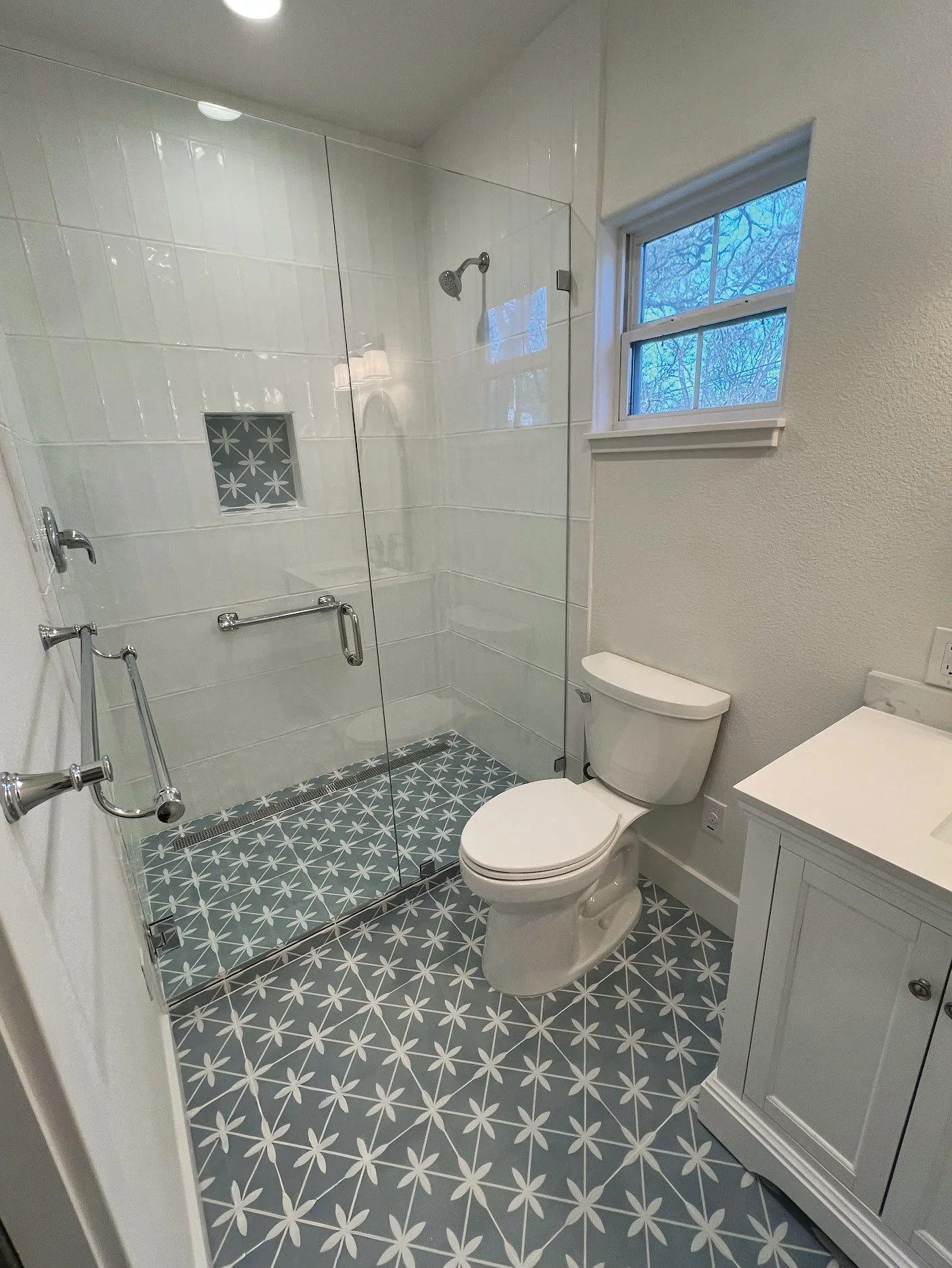 Bathroom with a glass shower enclosure, a white toilet, a window showing a view of trees, and patterned floor tiles with a star-like design.