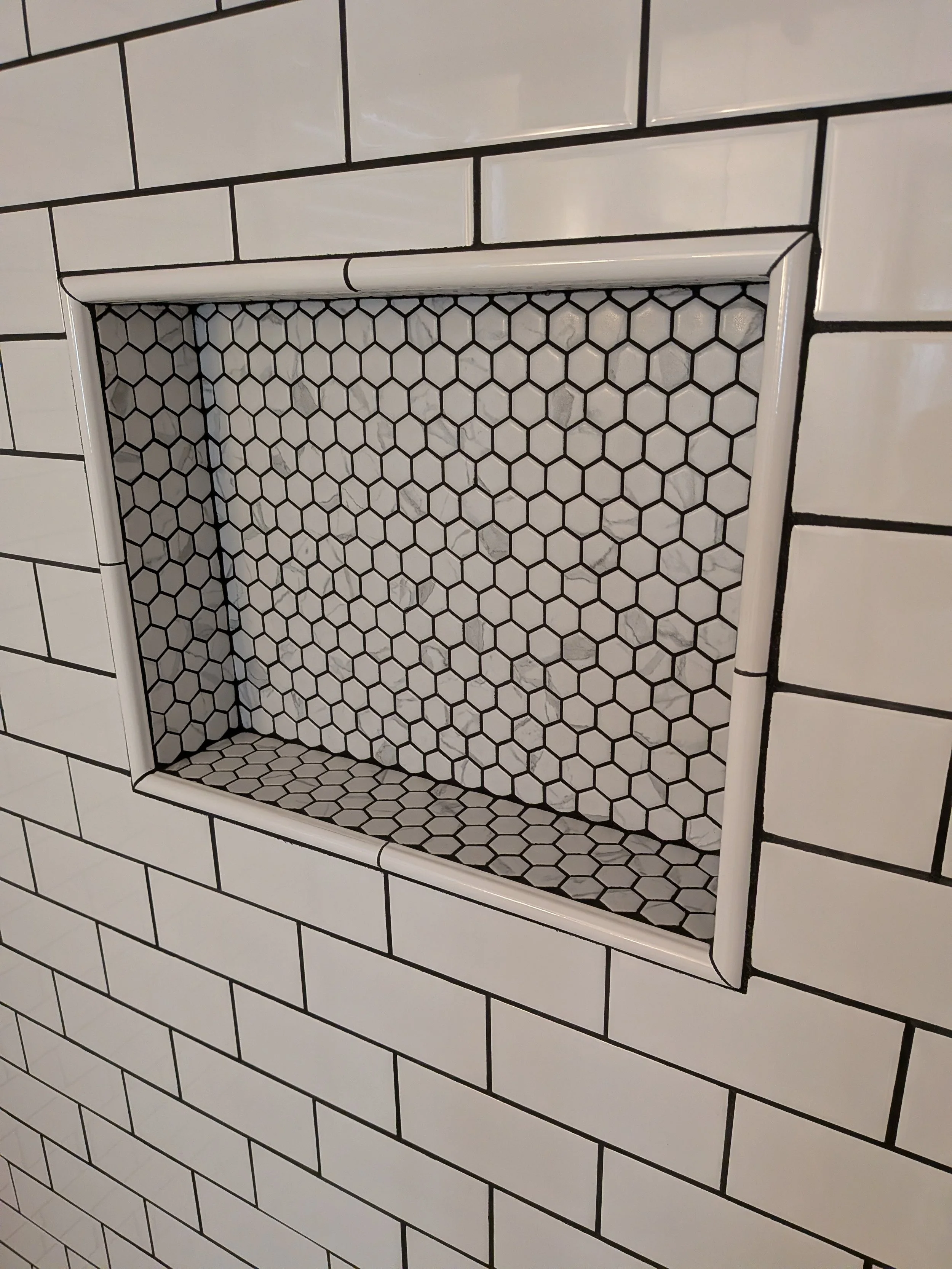 A decorative recessed shelf in a wall covered with white subway tiles with black grout lines. The shelf has a black hexagon tile pattern inside.