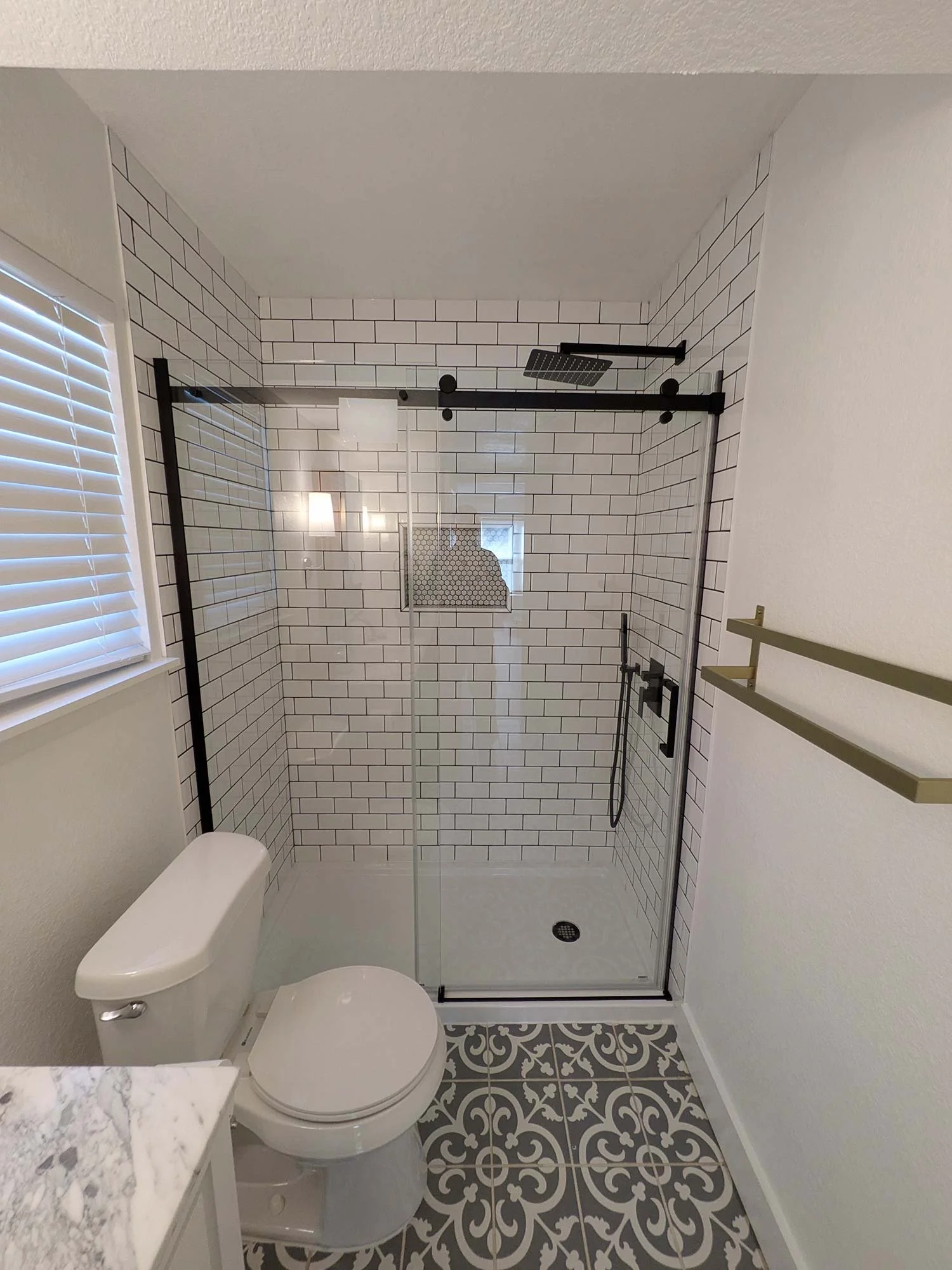Small bathroom with a walk-in shower with black metal framing, white subway tile walls, decorative black and white patterned floor tiles, a white toilet, a window with white blinds, and a small marble countertop.