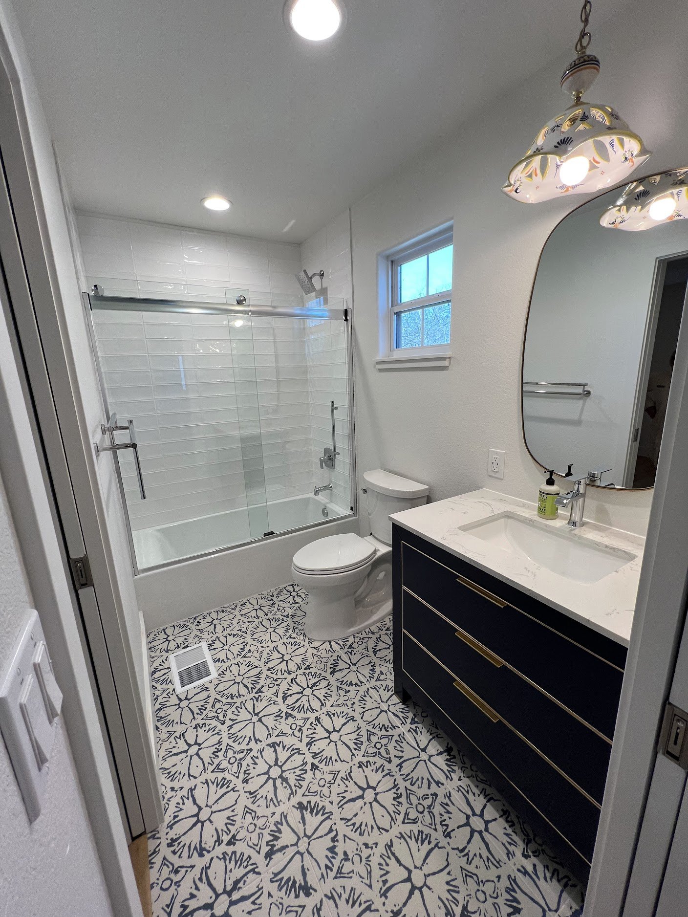 Bathroom featuring a walk-in shower with a glass door, toilet, vanity with a marble countertop, oval mirror, patterned tile floor, window with blinds, and ceiling lights.
