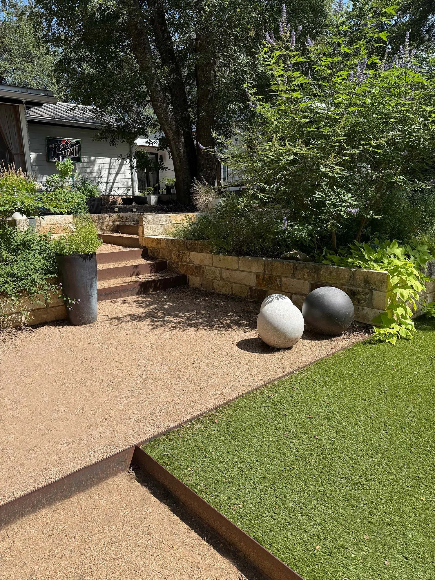 A garden scene with a sandy pathway, a potted plant, a stone retaining wall, lush green foliage, and a house in the background with a sign that reads 'PLASTIC'.