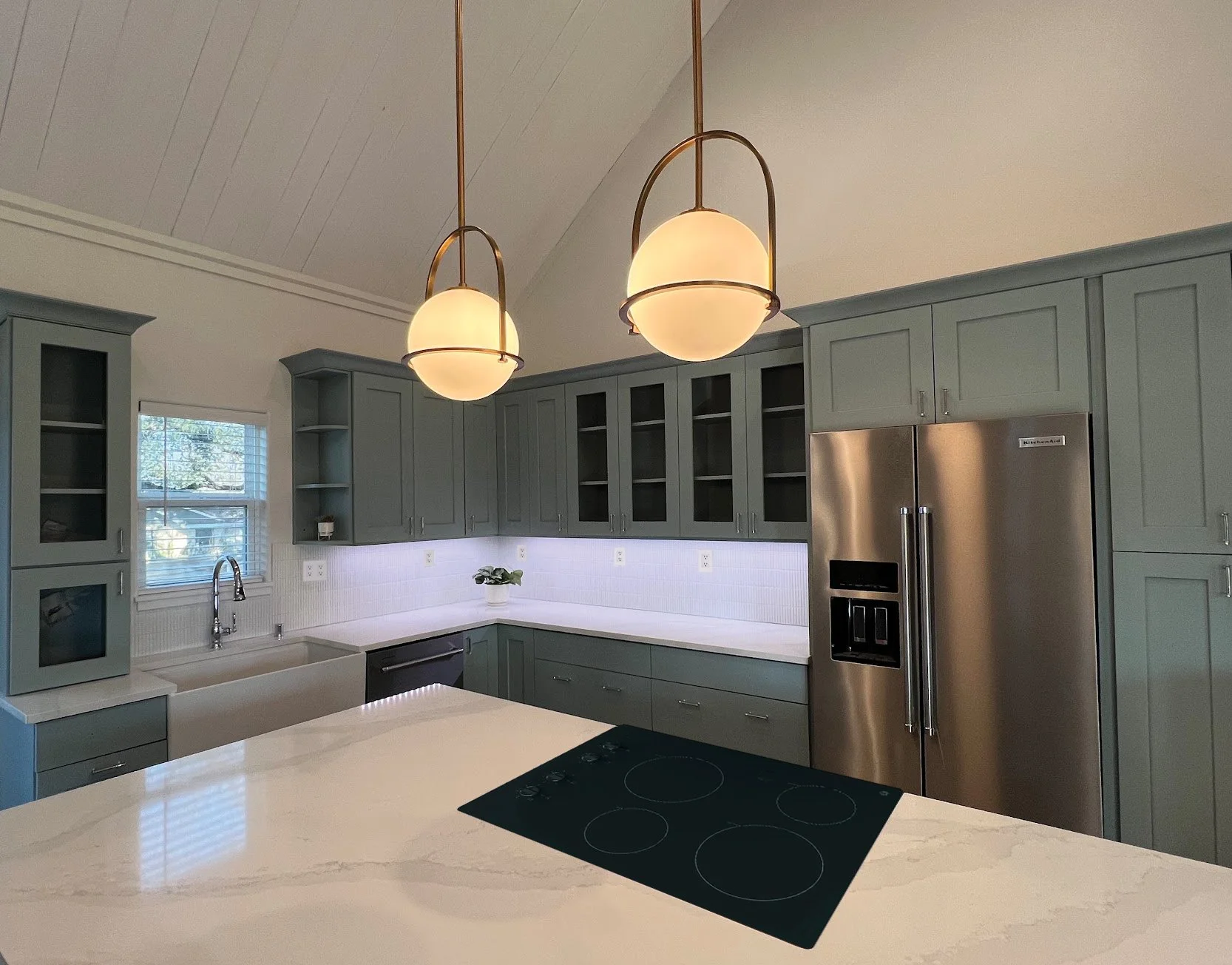 A modern kitchen with gray cabinetry, a white countertop, and stainless steel refrigerator. Pendant lights hang from the ceiling, and a window with blinds is above the sink. There is a potted plant on the counter.
