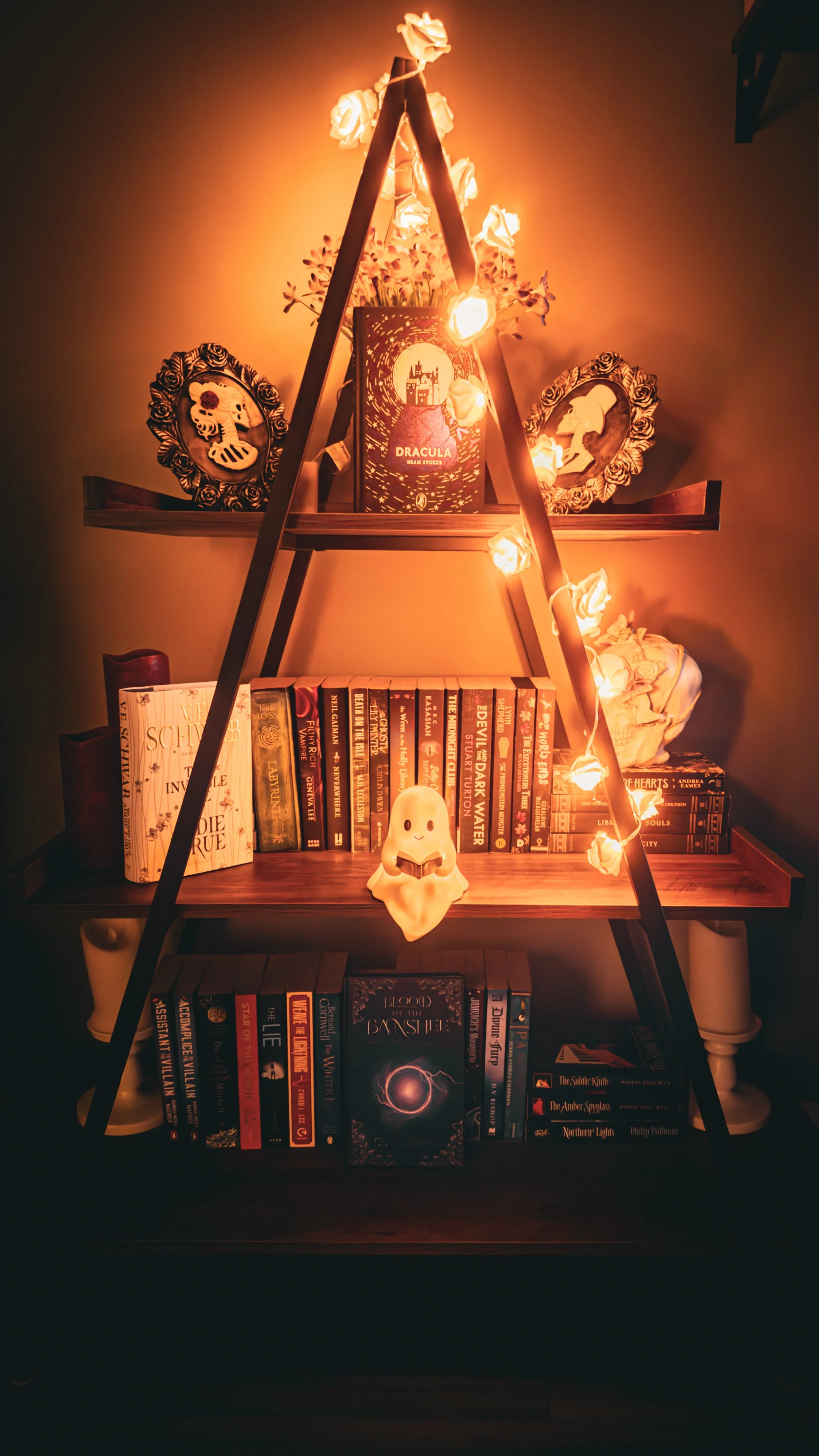 A wooden bookshelf with three shelves, illuminated by string lights, featuring books, decorative frames with skull and helmet silhouettes, a small ghost figurine, and a Dracula book in the middle shelf.