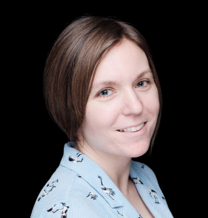 A woman with short brown hair and light skin, smiling at the camera, wearing a light blue shirt with a dark pattern, against a black background.
