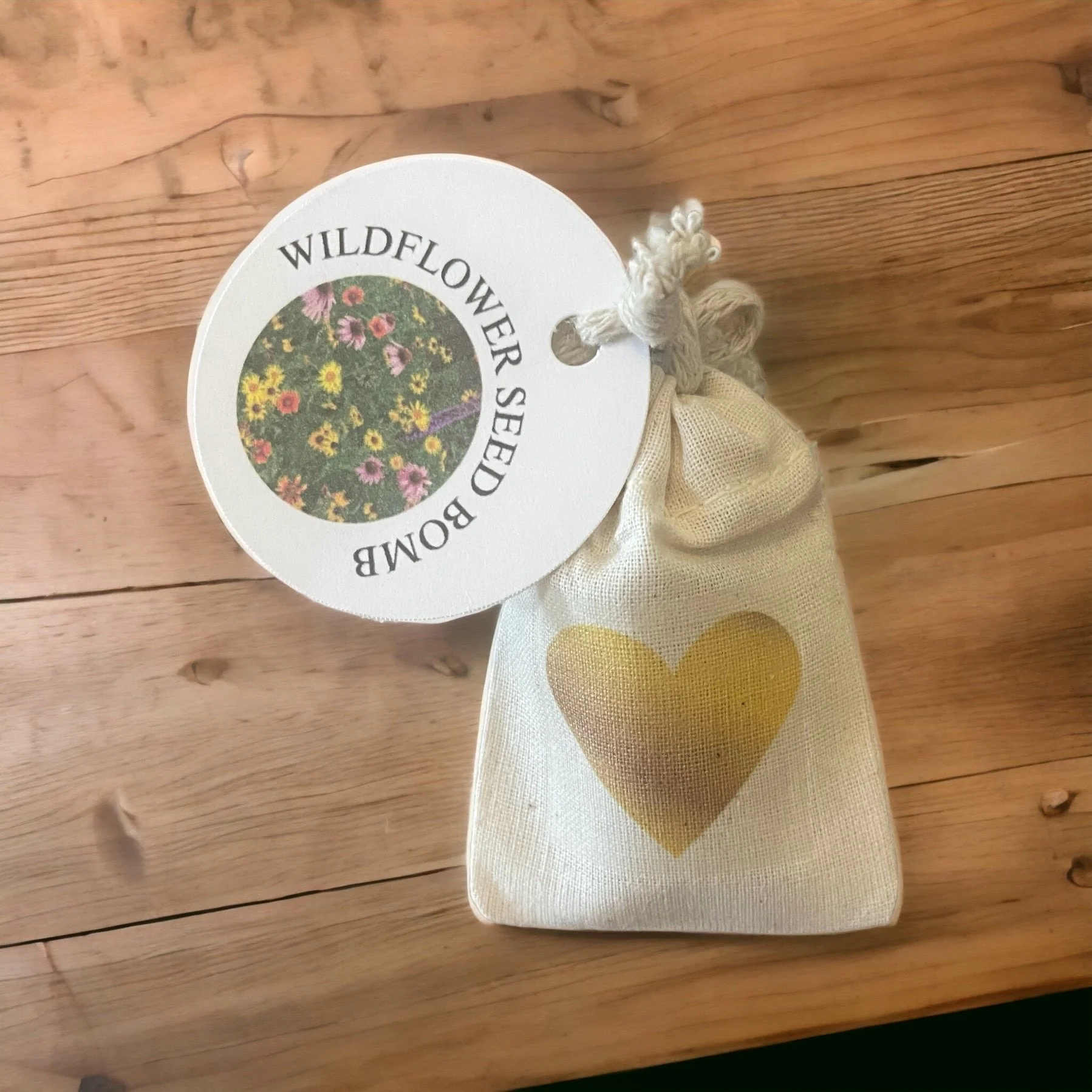 A round white tag with the words 'Wildflower Seed Bomb' and a colorful photo of wildflowers in a circular frame, attached to a beige cloth bag with a gold heart design.