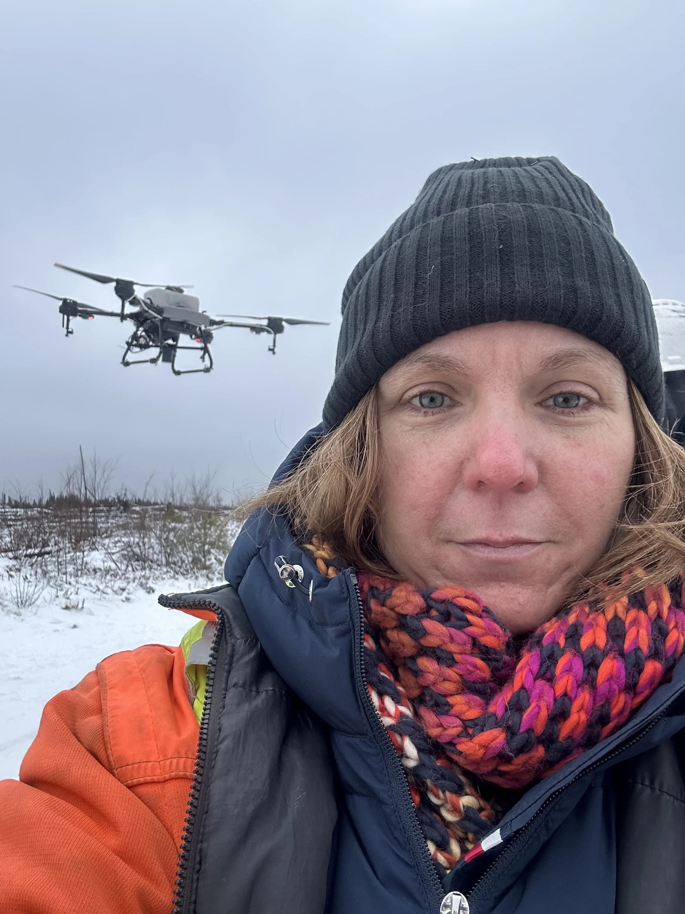 A woman with light skin and blonde hair wearing a black beanie and colorful scarf, dressed in outdoor winter clothing, with a drone flying in the cloudy sky behind her in a snowy landscape.