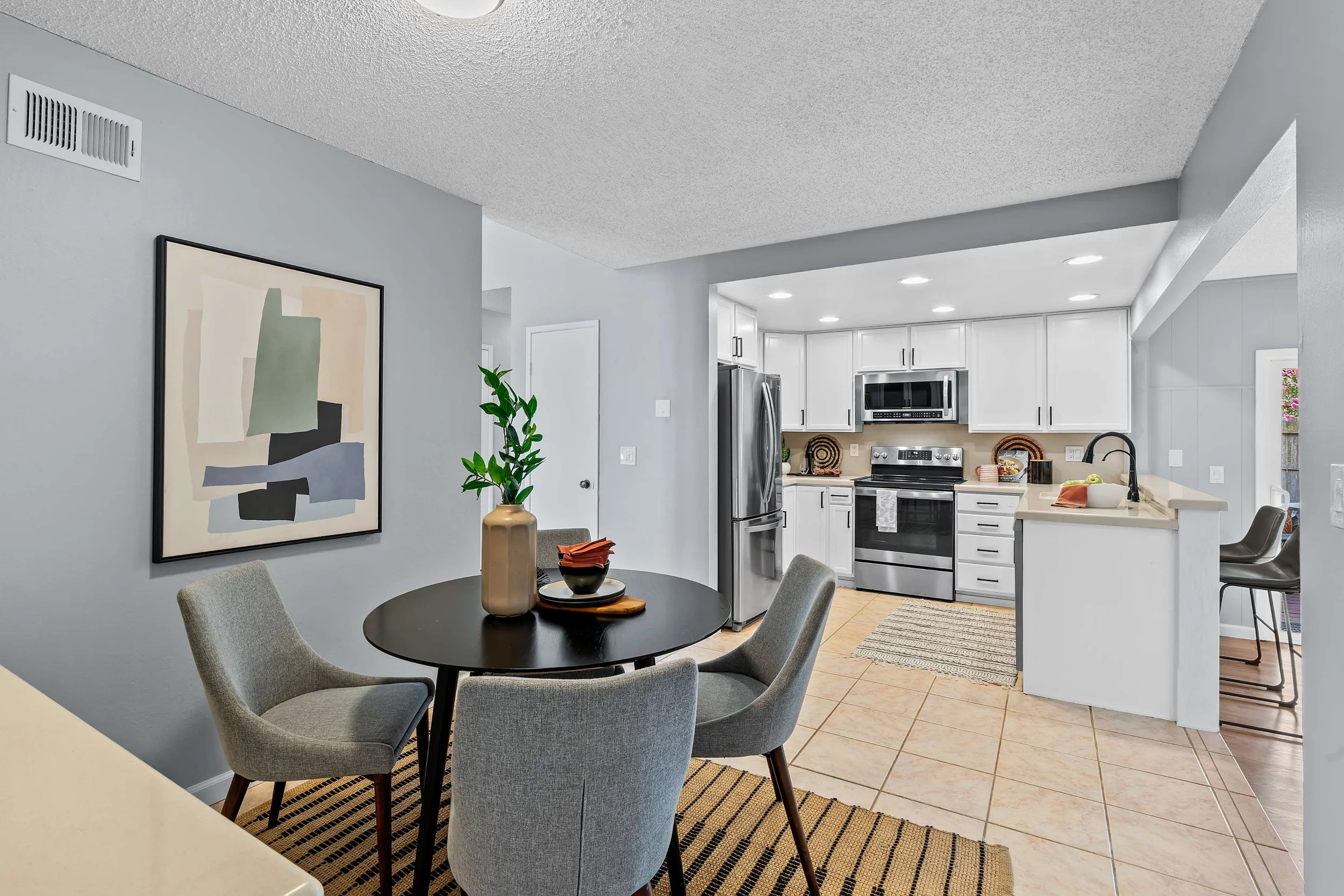Open-concept kitchen and dining area with a round black table, four gray upholstered chairs, and modern white cabinetry. The kitchen features stainless steel appliances and a microwave, with recessed ceiling lights. A decorative art piece hangs on the light gray wall, and a large tan vase with a green plant is on a side table. The floor is tiled, with a striped rug near the kitchen island.