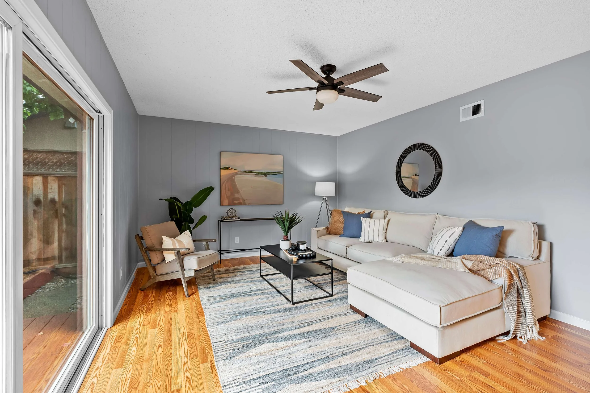 Living room with a beige sofa, blue and beige throw pillows, wooden floor, glass sliding door to outside, artwork of a beach scene on the wall, potted plant, side table with decor, black coffee table, striped area rug, ceiling fan with wooden blades.
