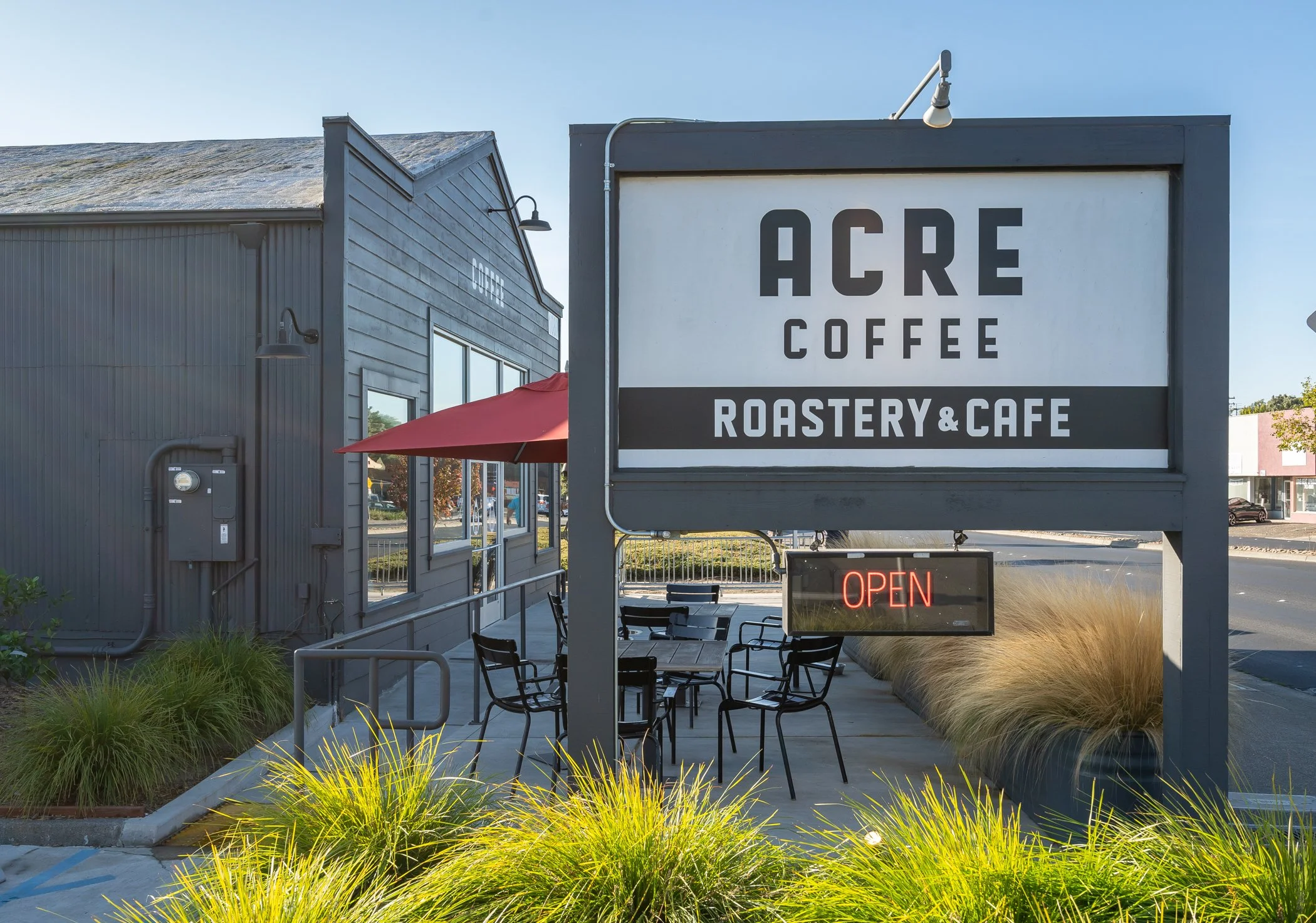 Outdoor cafe with a large sign monolith reading 'Acre Coffee Roastery & Cafe', with a small lit 'Open' sign underneath. There are black metal chairs and tables outside, and a red umbrella is visible near the entrance. The building has gray siding wit