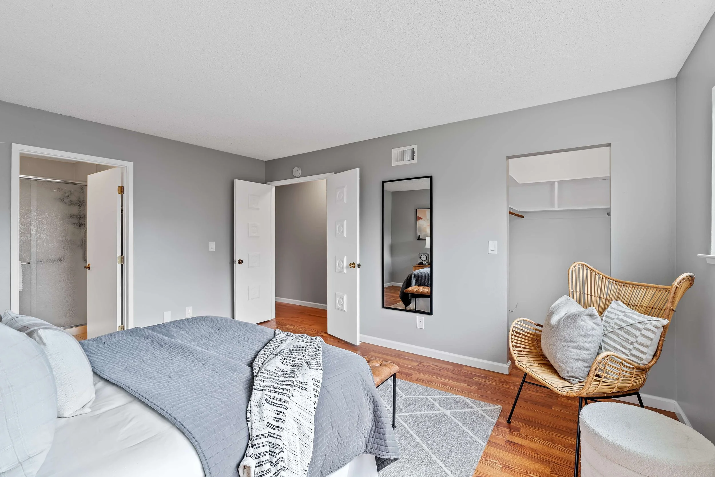 A bedroom with a bed, a mirror, a wicker chair with pillows, open closet doors, and a bathroom door in the background.