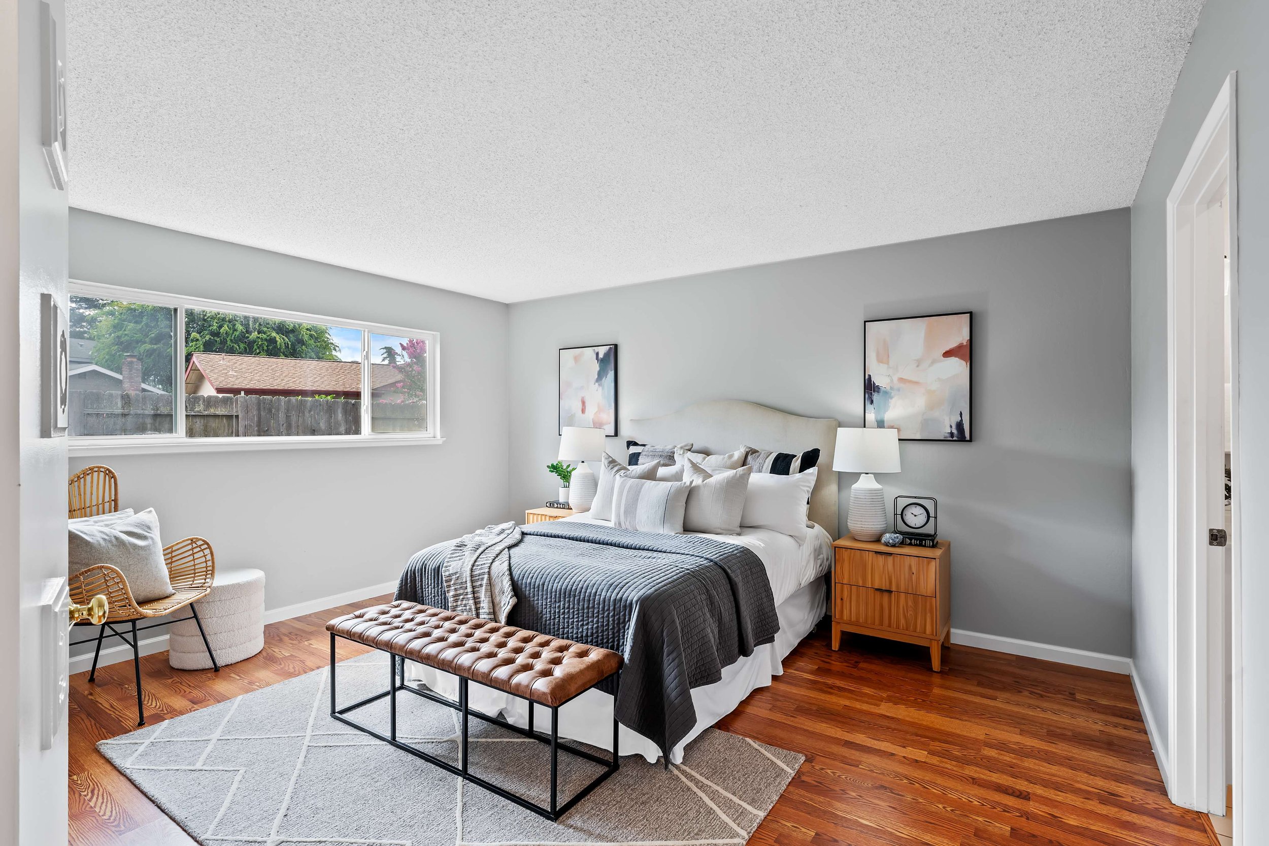 A cozy bedroom with a large bed, framed abstract art on the wall, wooden nightstand with a lamp and clock, a window showing a fenced backyard, a wicker chair, and a light gray rug on hardwood floor.