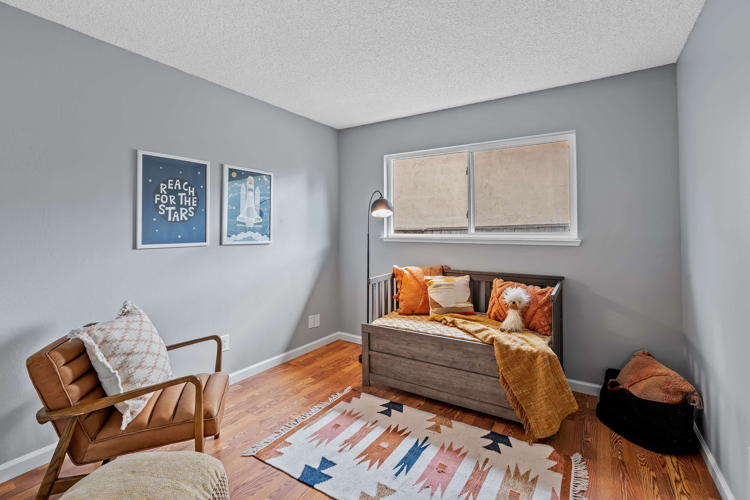 A cozy corner of a room with a wooden chair with a patterned cushion, a small wooden bed with orange and beige pillows and a blanket, a black basket with a dog, and framed space-themed art posters on the light gray wall.