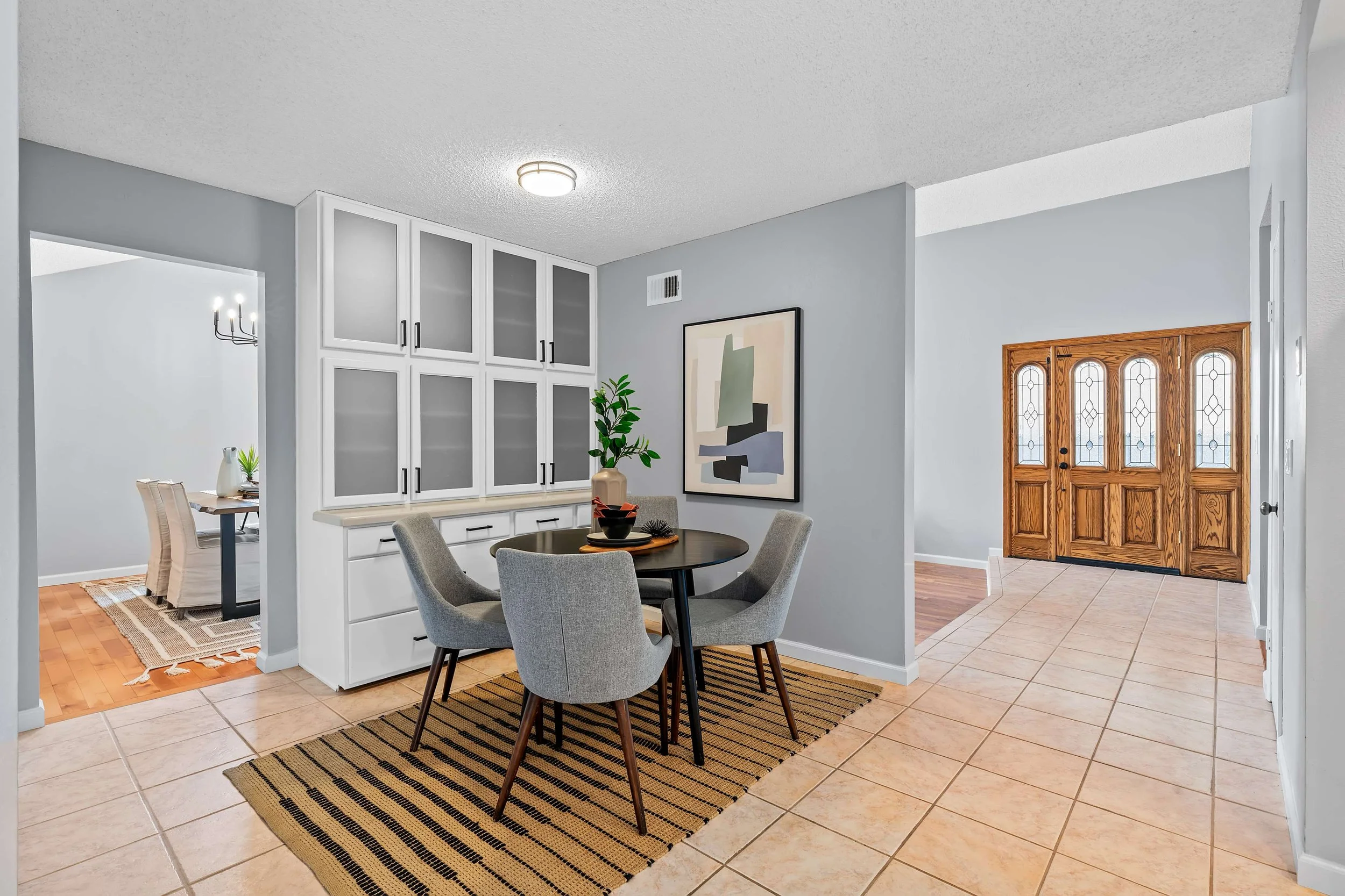View of a dining area with a round table and four grey upholstered chairs, a stairway, a white built-in cabinet, and artwork on the wall. The floor has beige tiles and a black-striped rug, with a front door featuring wood and glass designs visible in