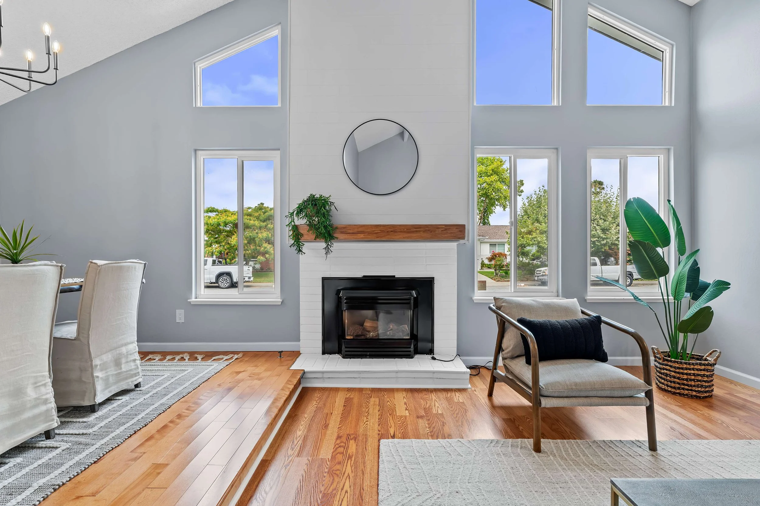 A modern living room with a white fireplace, large multi-pane windows showing a neighborhood, light wood floors, a plant in a woven basket, and a comfortable armchair with a cushion.