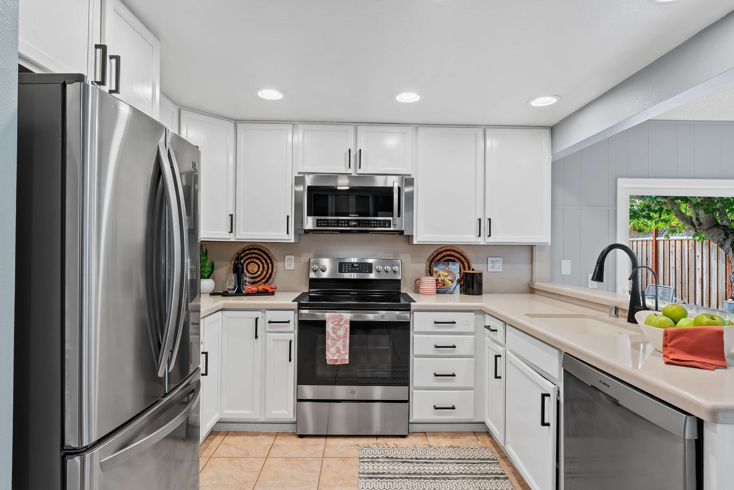 Modern kitchen with white cabinets, stainless steel appliances, and a window showing a view of a tree and a wooden fence outside.