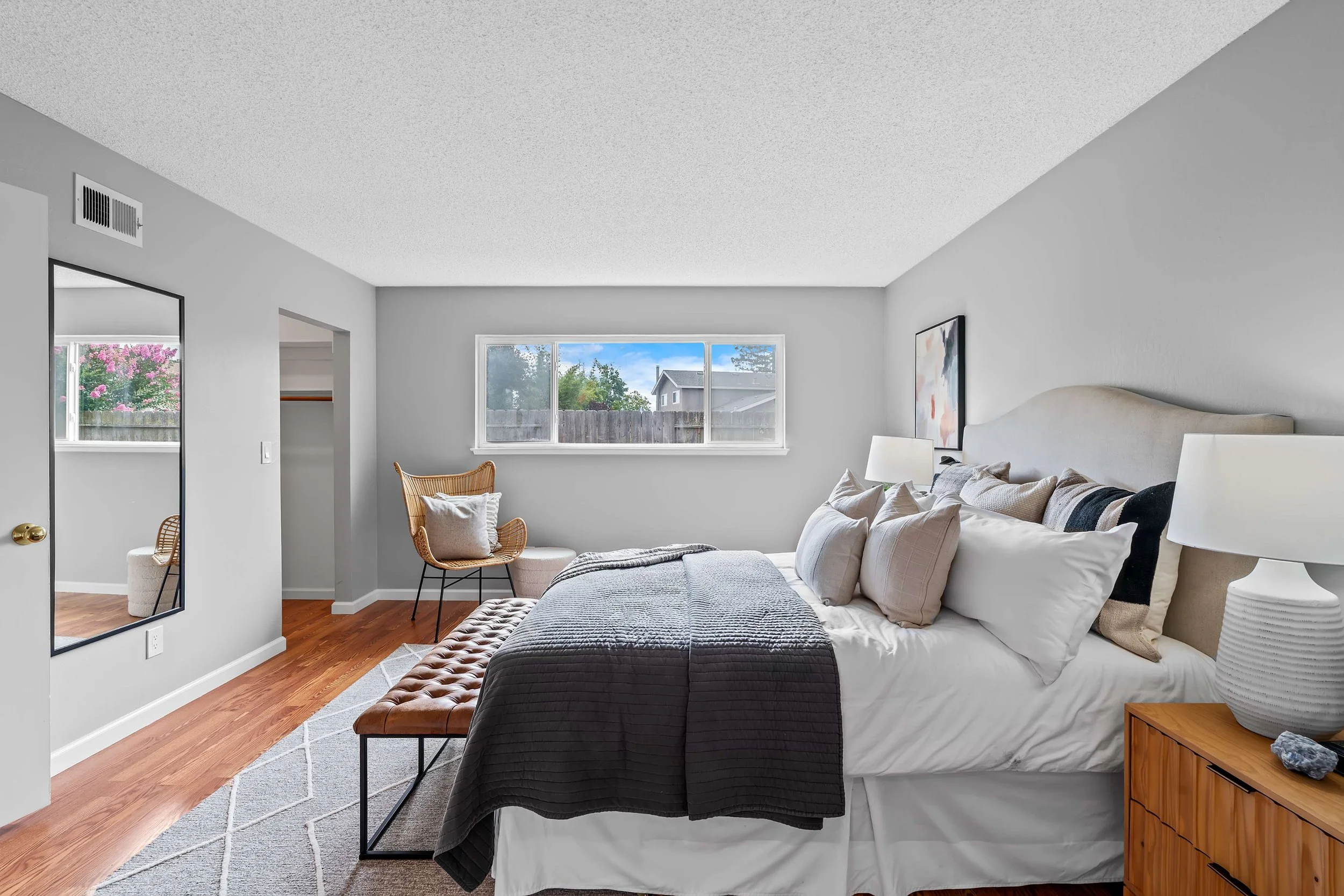 A bedroom with a large bed, nightstand, and window showing a backyard with trees and sky.