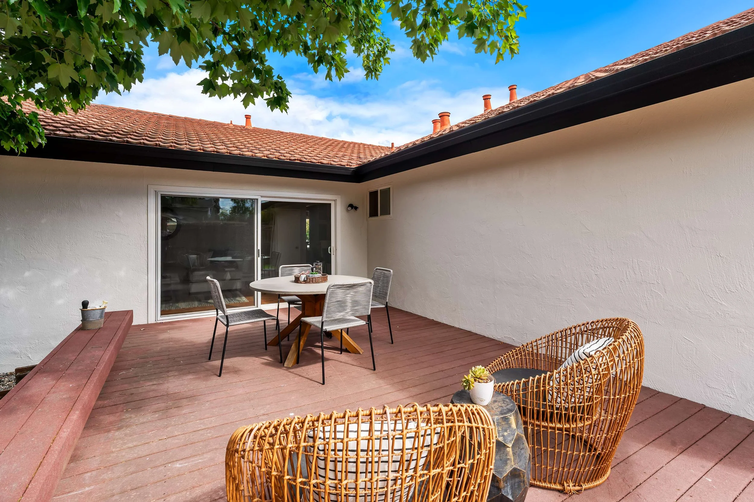 Outdoor patio area with a round table and four chairs, a woven wicker chair, potted plants, a sliding glass door, a red wooden bench, and a white stucco wall, with a tiled roof and a tree with green leaves overhead.