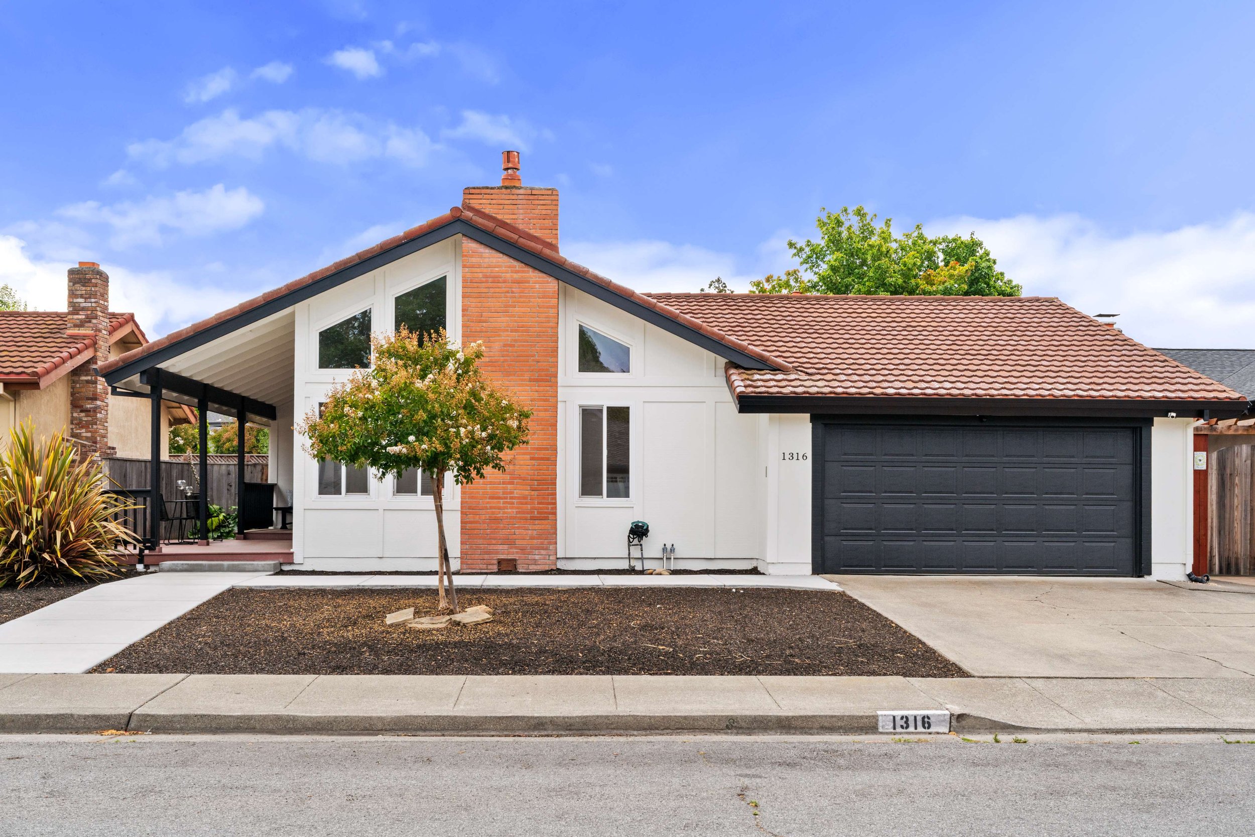 Modern residential house with brick and white exterior, black garage door, small front yard with young trees, and a sidewalk. Clear sky with a few clouds.