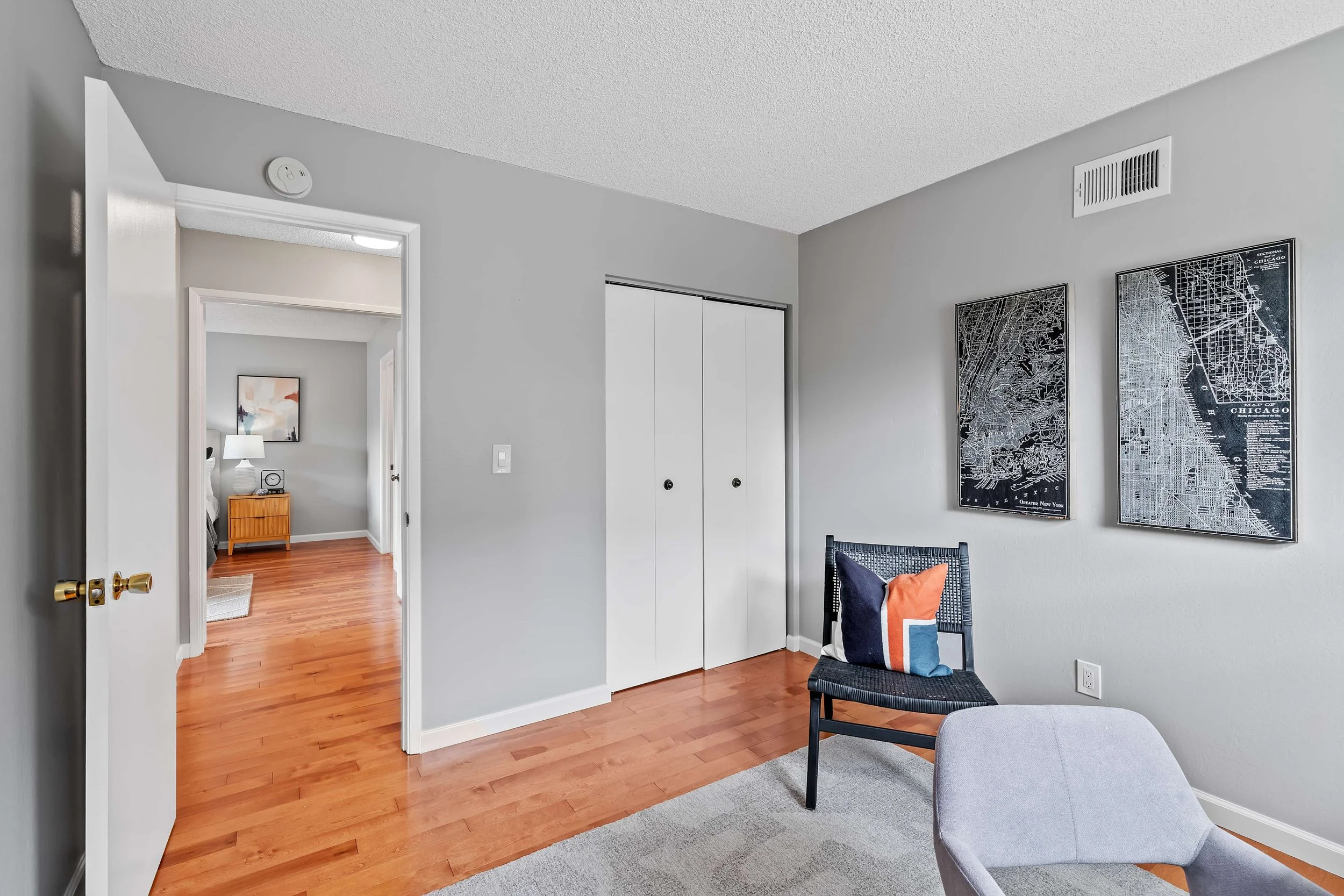 A corner of a room with a gray wall, wooden floor, and a white door. Two black-framed maps of Chicago are hanging on the wall, and a black chair with a pillow is placed beside a light gray upholstered piece of furniture.