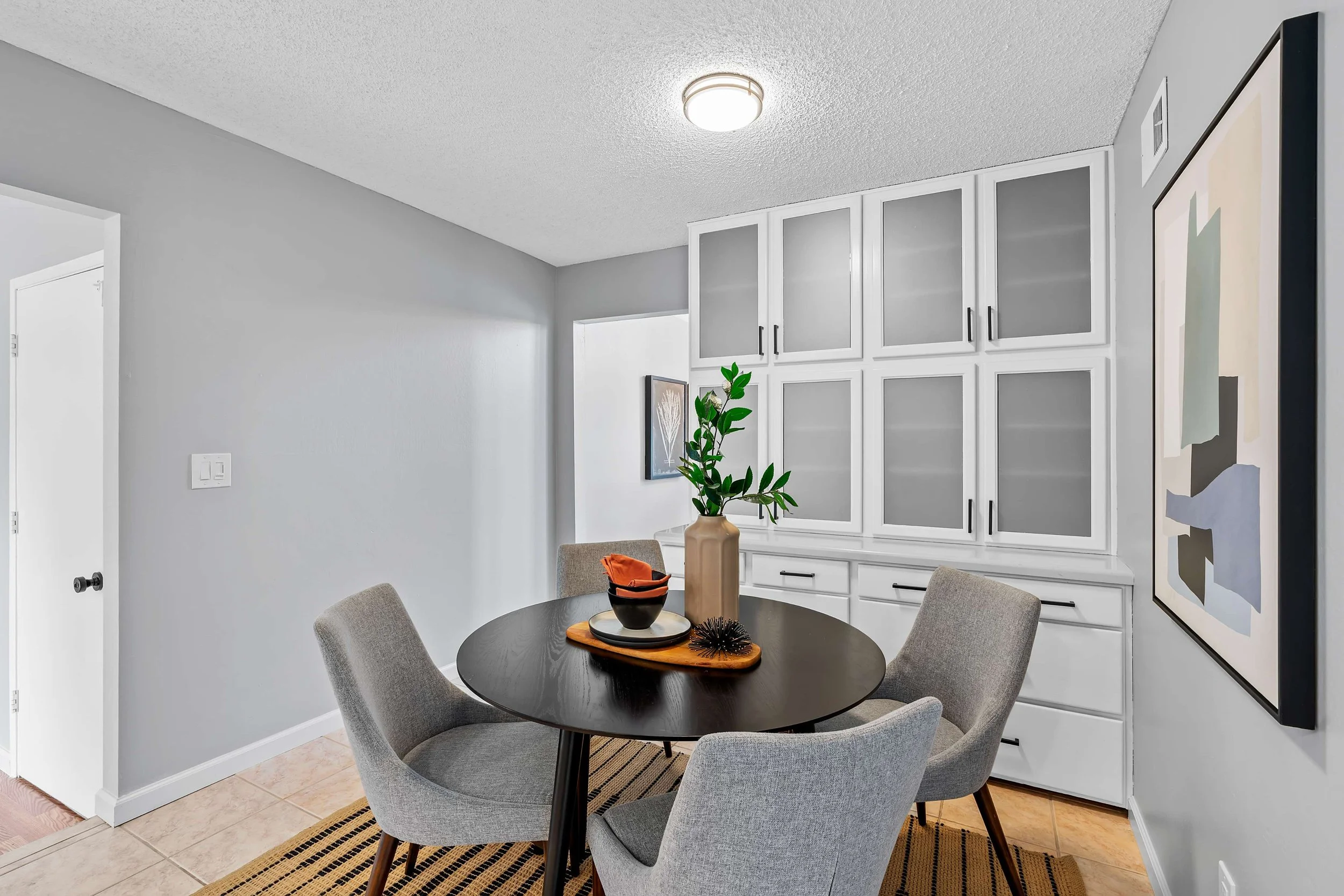 A dining room with a black round table and four light gray upholstered chairs. There is a vase with green leaves and a tray with decorative items on the table. White cabinets with glass doors are on the wall, and a piece of artwork is partially visib