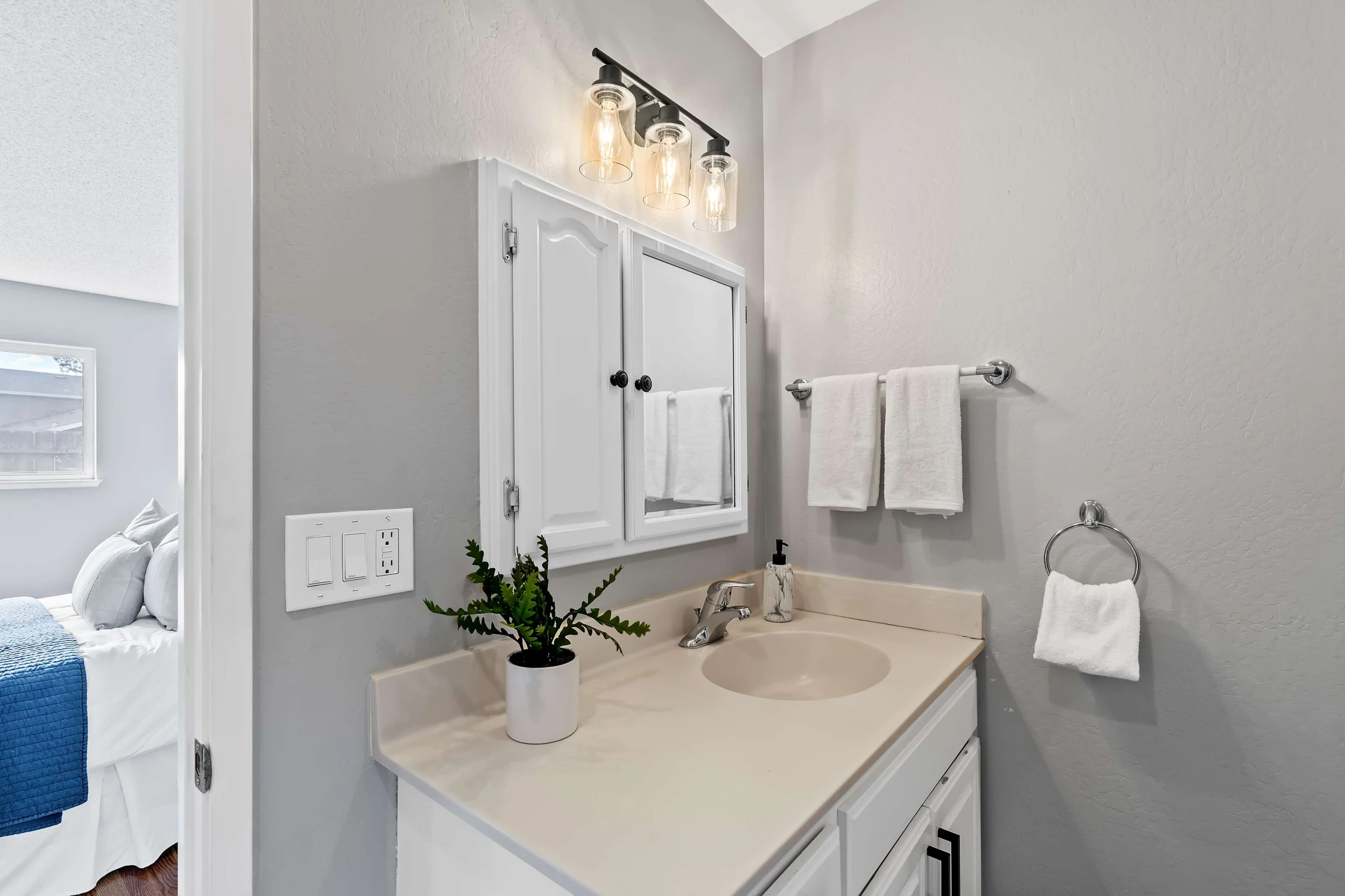Bathroom vanity area with a beige countertop, a white cabinet, a mirror, a silver faucet, a small potted plant, a soap dispenser, and towels hanging on a silver towel bar. Wall lights illuminate the space, and a window is partially visible on the lef