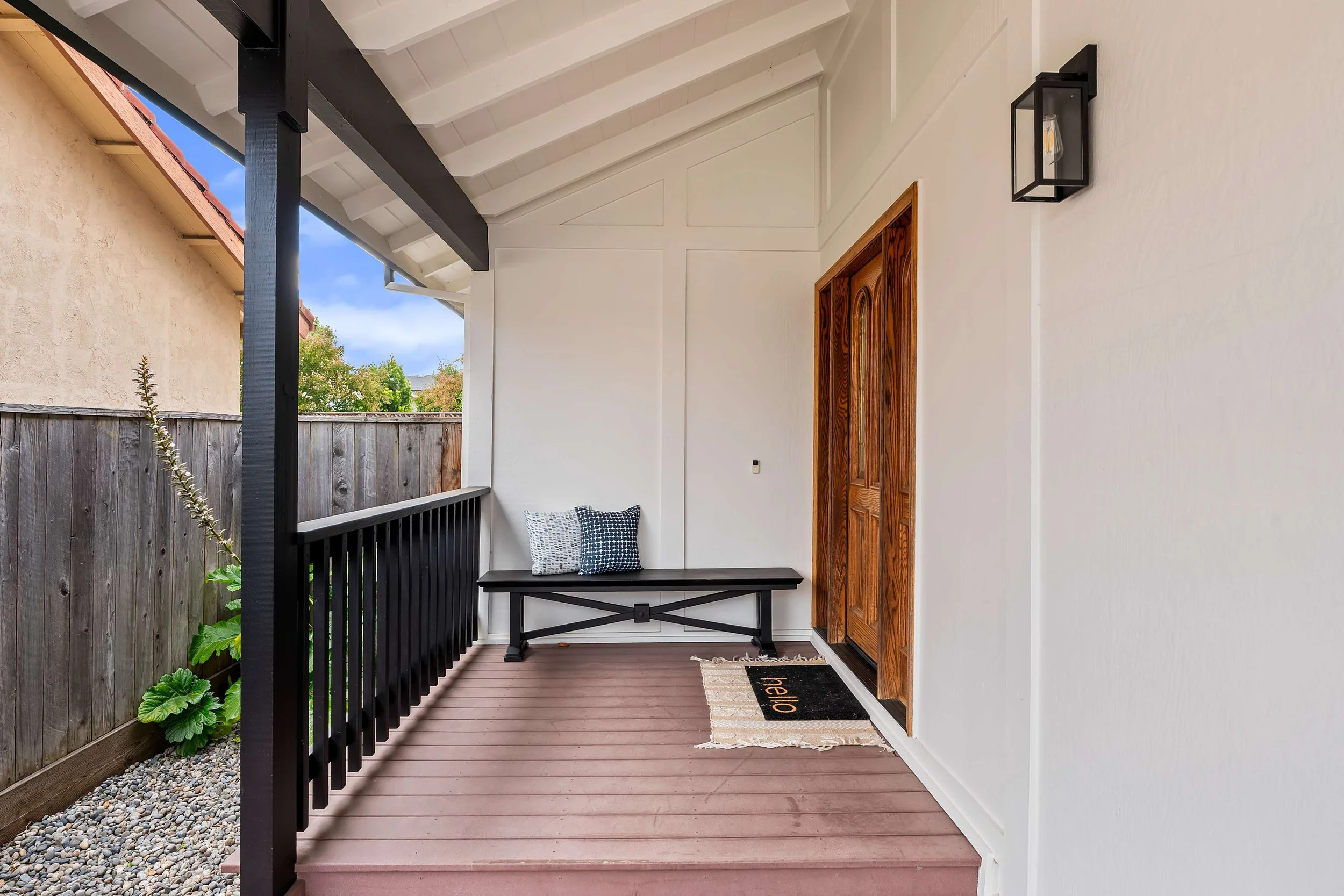 Front porch area with a wooden door, black bench with patterned cushions, black outdoor wall lantern, white wall, and brown deck flooring.