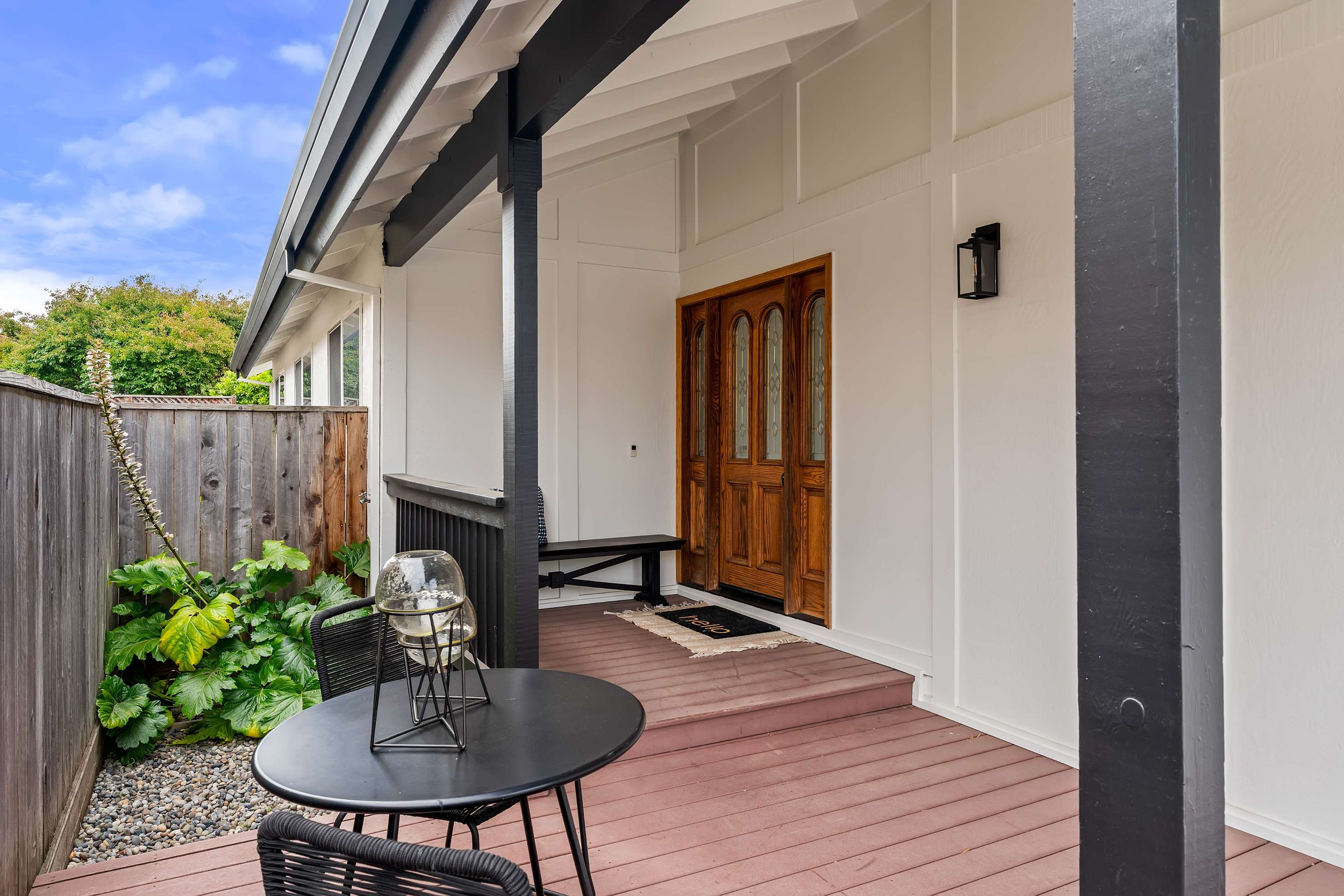 A cozy porch area with a black round table and chairs, a wooden front door with glass insets, a black outdoor wall lantern, a hanging rug at the door, and lush green plants by a wooden fence, under a blue sky with white clouds.