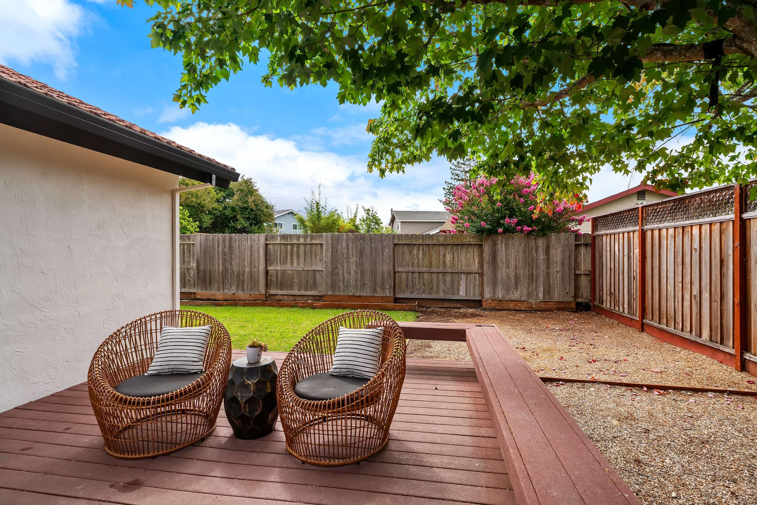 A backyard patio area with three wicker chairs with black cushions and striped pillows around a small round table with a potted plant. The patio is adjacent to a house with a white wall, and a tall wooden fence surrounds the yard. There are green tre