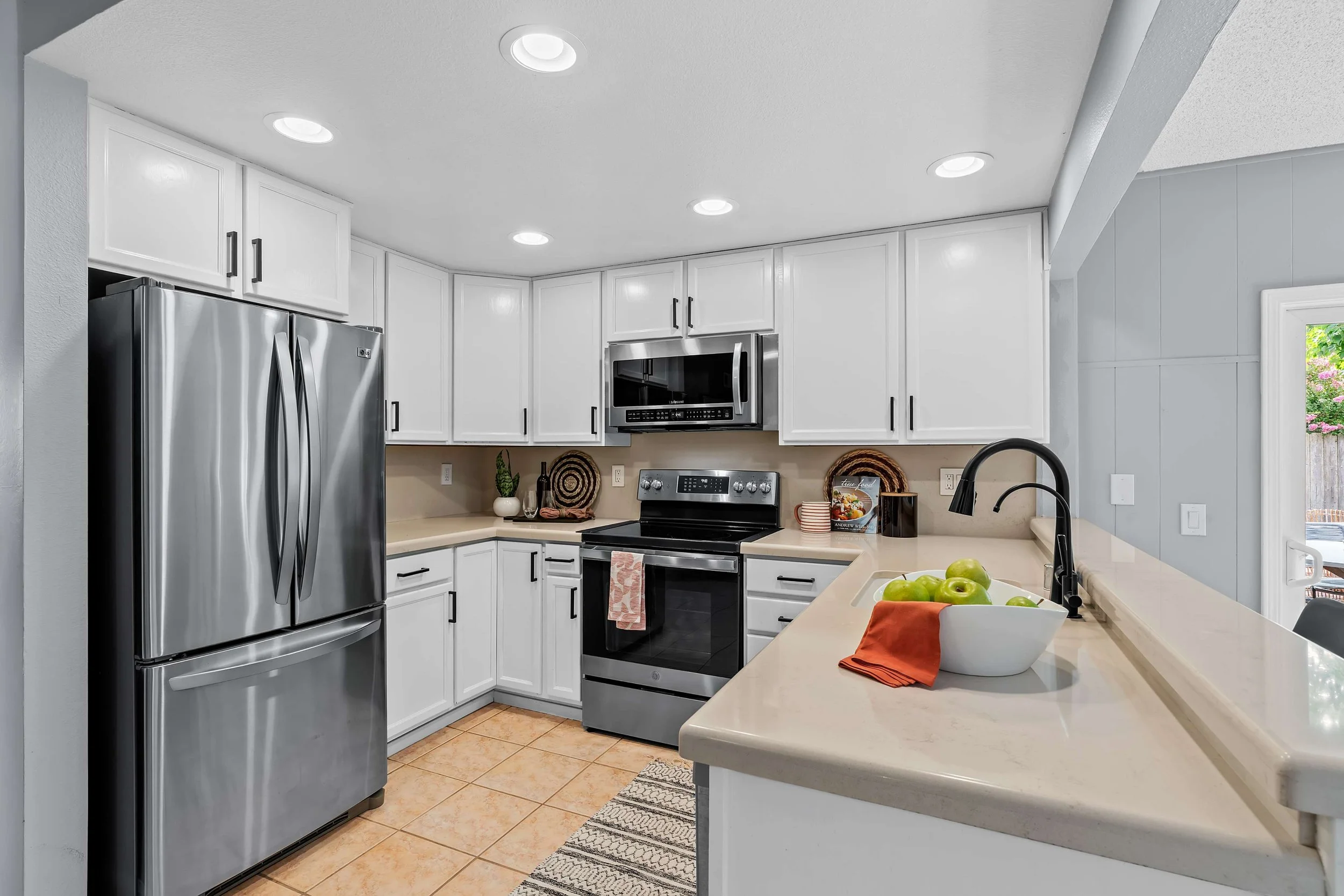 A modern kitchen with white cabinets, stainless steel refrigerator and microwave, beige countertops, a black stovetop and oven, a bowl of green apples, orange napkin, black faucet, and small decorative items on the counter. Recessed lighting in the c