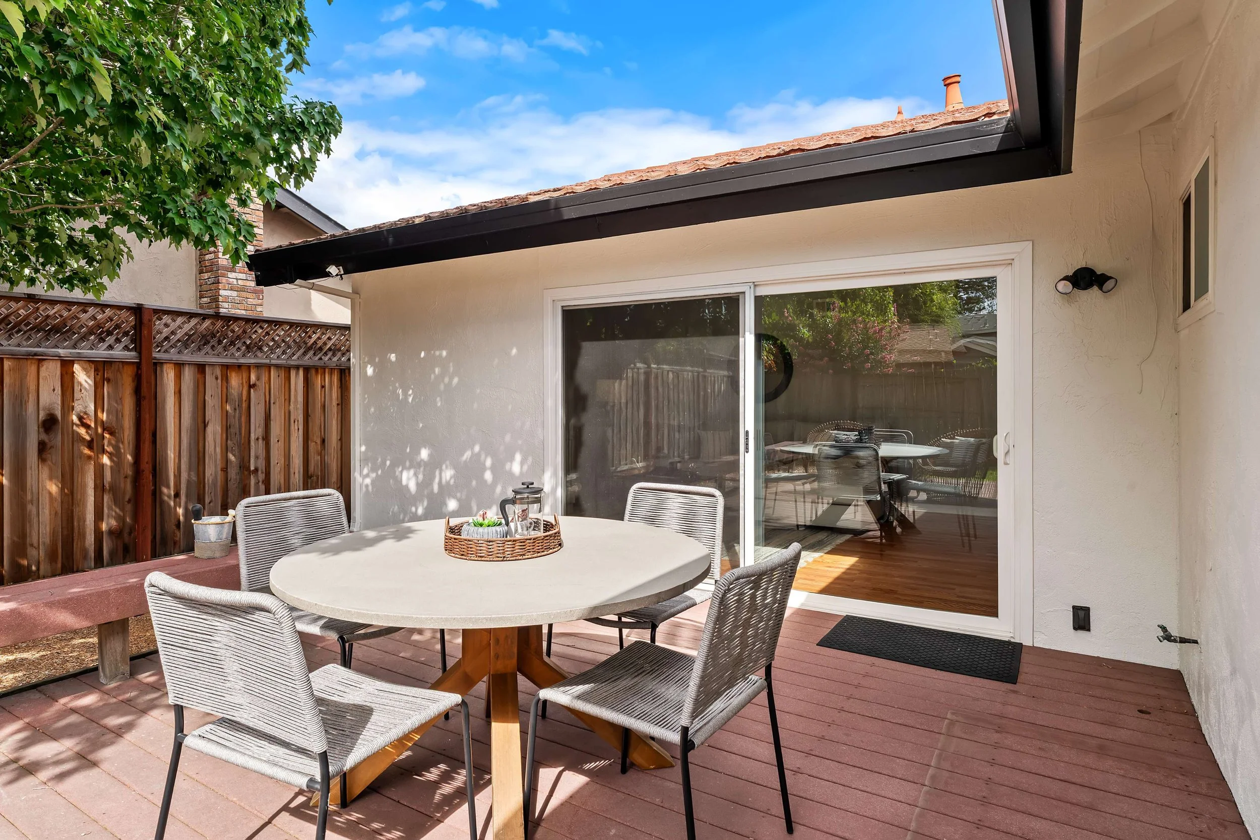 An outdoor patio area with a round table, four chairs, and a small tray with a pitcher and cups, surrounded by a wood fence and a tree, with a sliding glass door leading to an indoor space, under a cloudy blue sky.