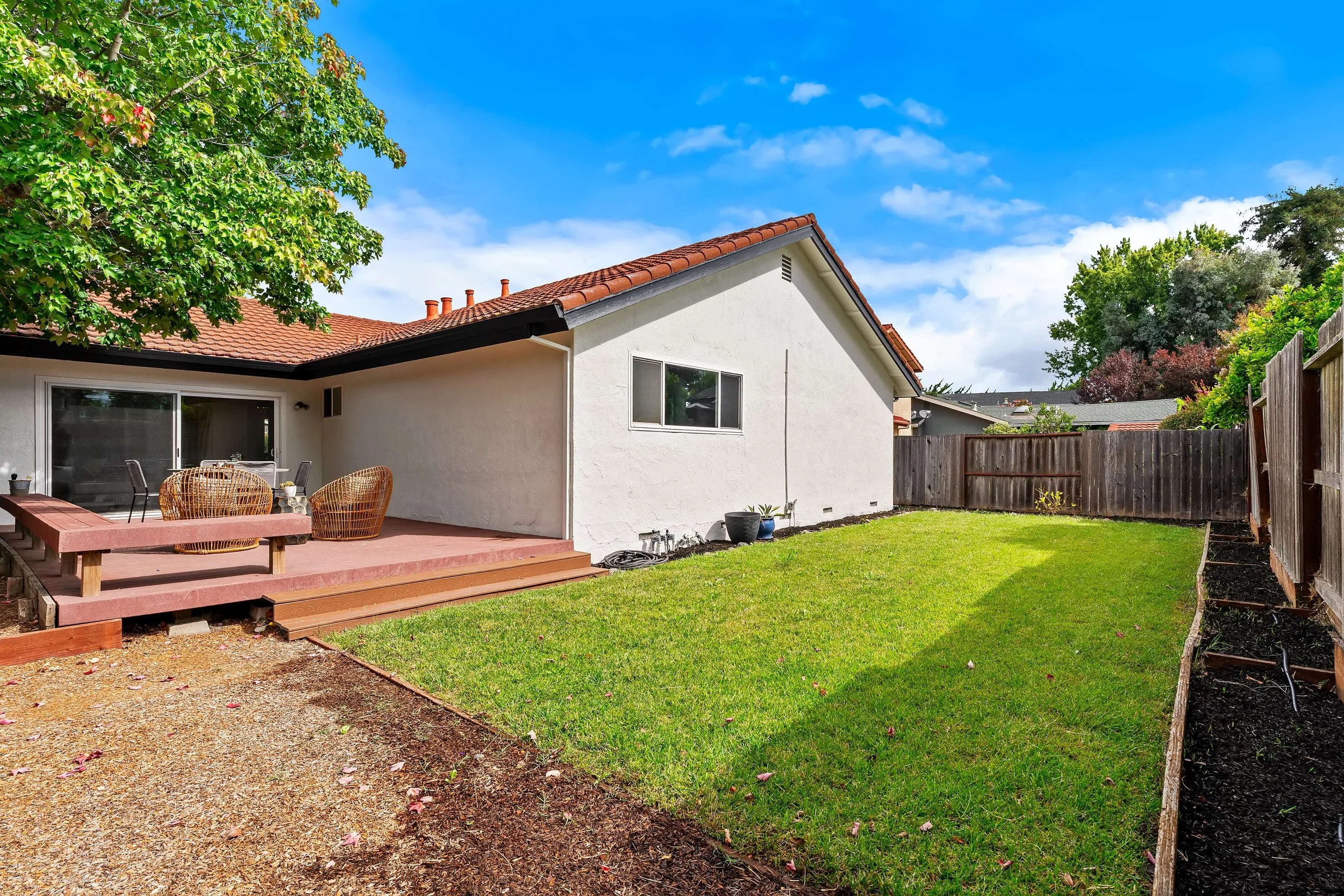 Backyard with patio, outdoor table and chairs, green lawn, trees, and a wooden fence under a partly cloudy sky.