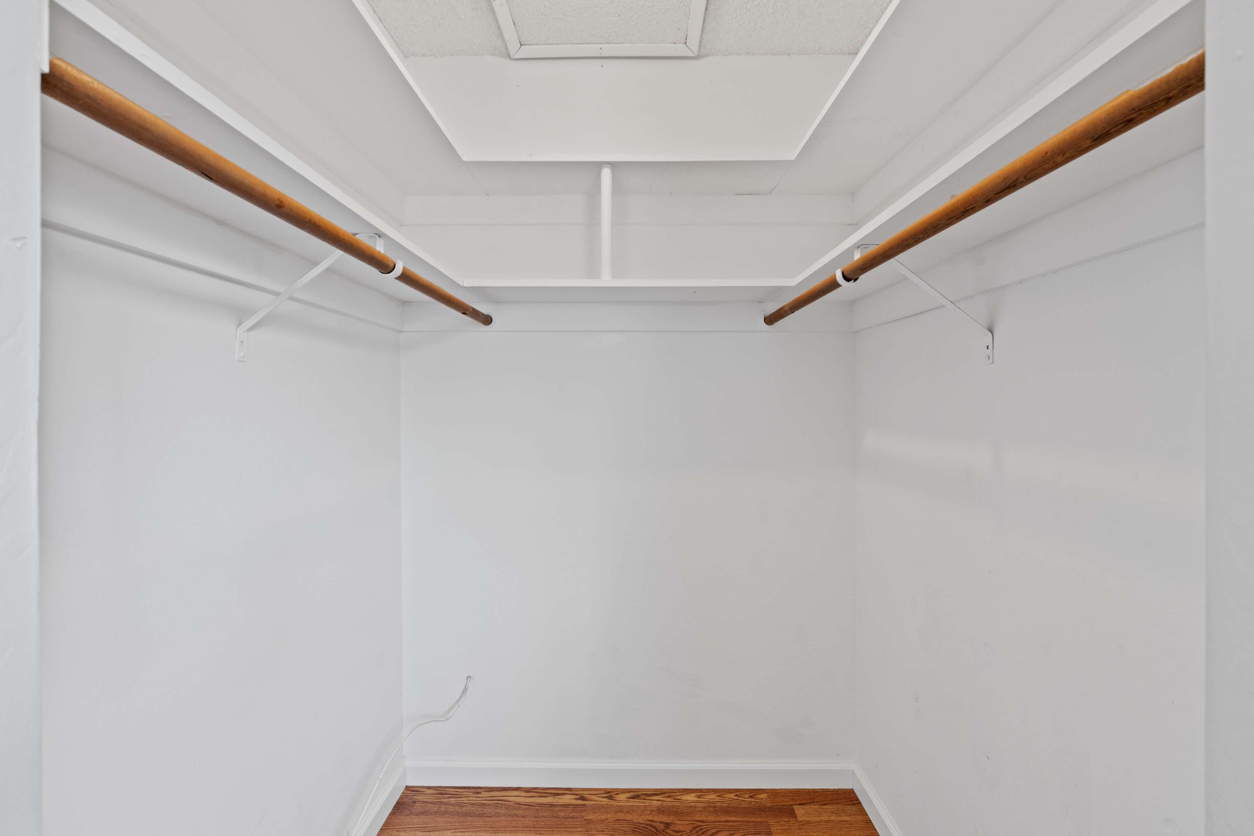 Empty walk-in closet with white walls, two brown hanging rods, a ceiling vent, and wooden flooring.