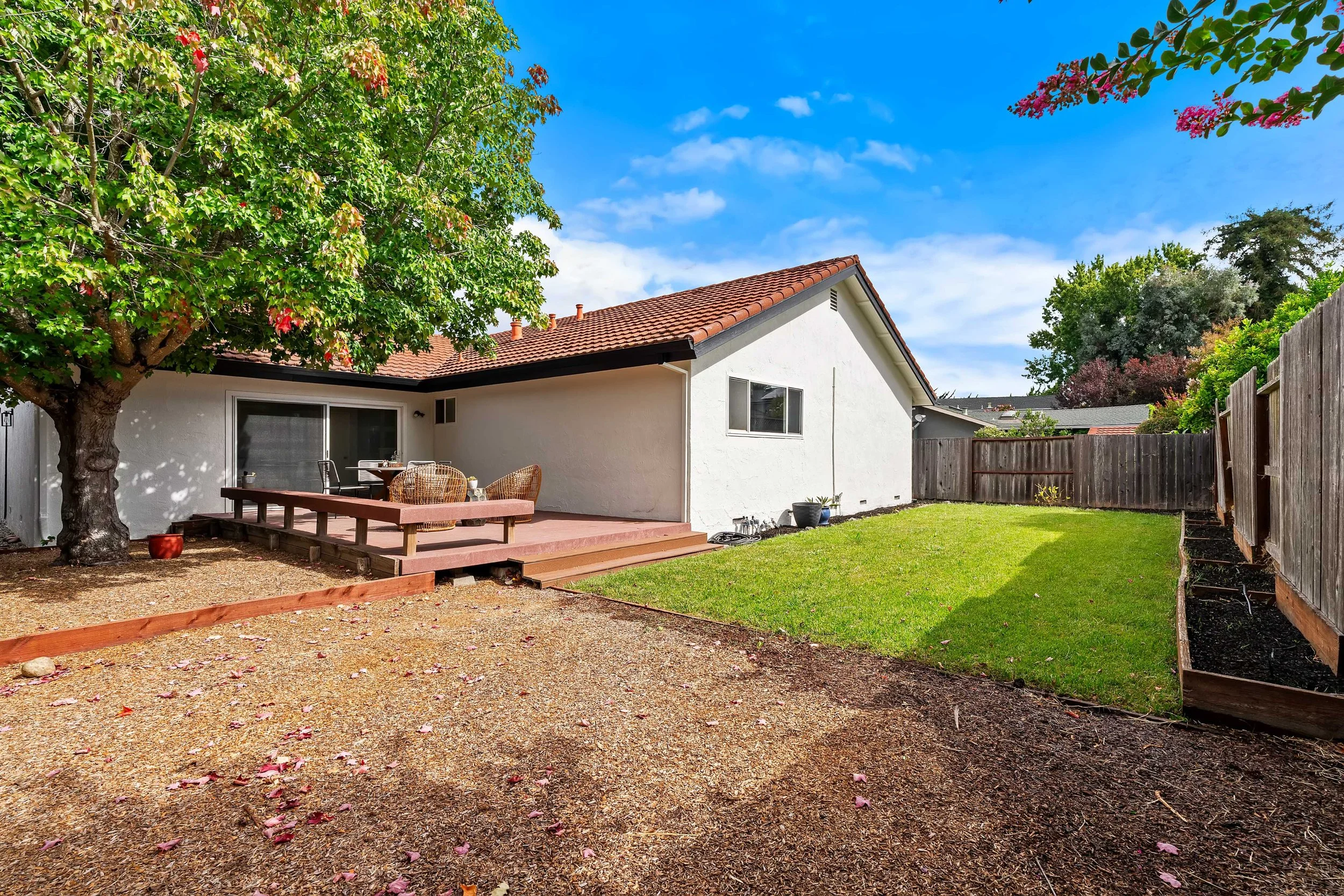 Backyard of a house with a patio, a large tree with green leaves, a wooden fence, and a green lawn under a blue sky with some clouds.
