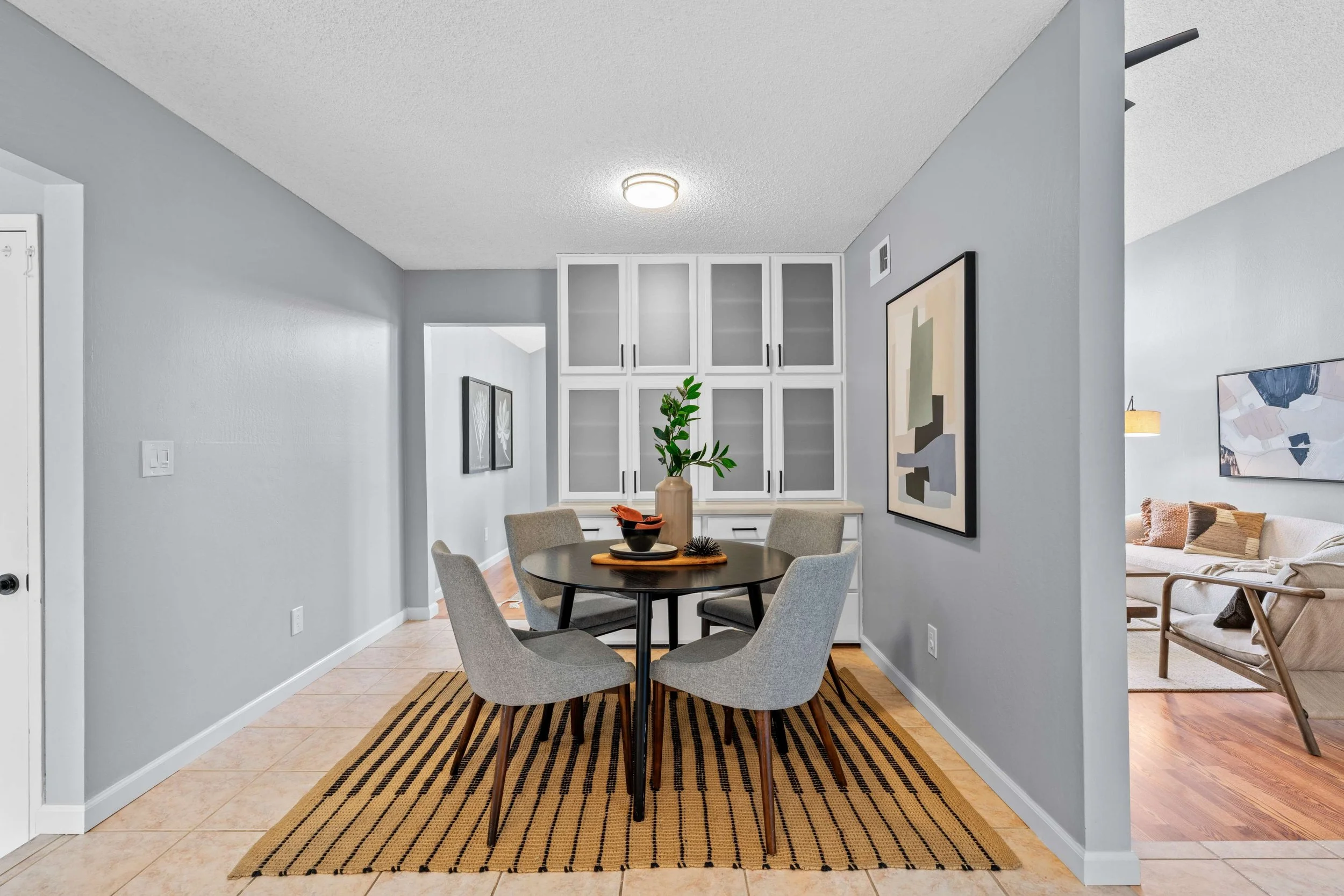 Interior view of a dining area with a round black table, six light gray upholstered chairs, a beige and black striped rug, a beige vase with green plants, against light gray walls, in a modern home.