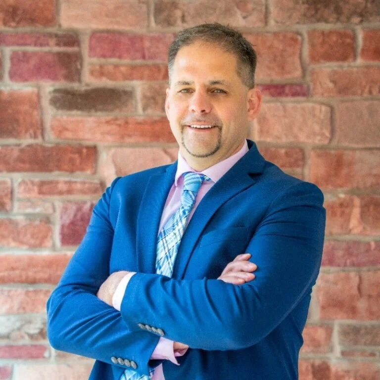 A man in a blue suit with a patterned tie, light pink shirt, and crossed arms standing in front of a brick wall.