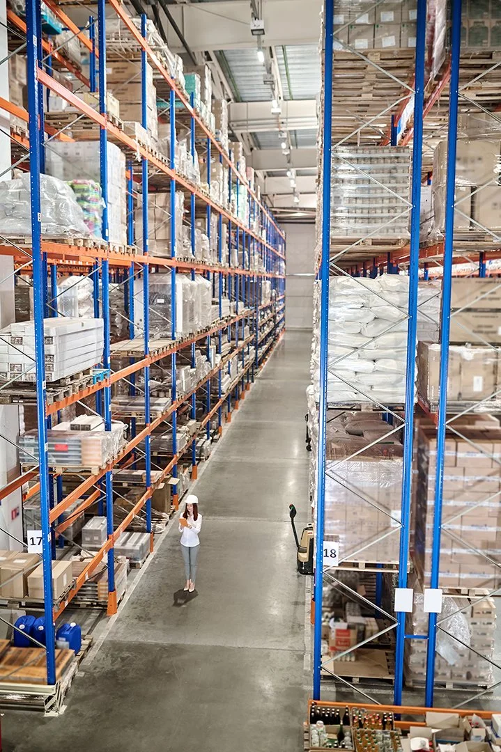 A woman in a warehouse aisle wearing a white safety helmet and white top, standing near shelves stocked with boxed and packed items.