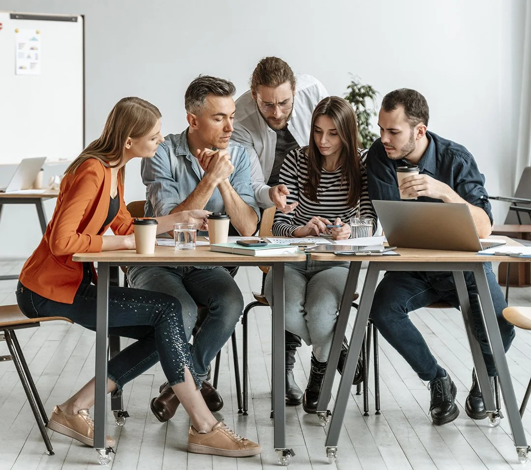 Group of five young adults working together in a meeting room, gathered around a table with laptops, papers, and coffee cups, discussing a project.