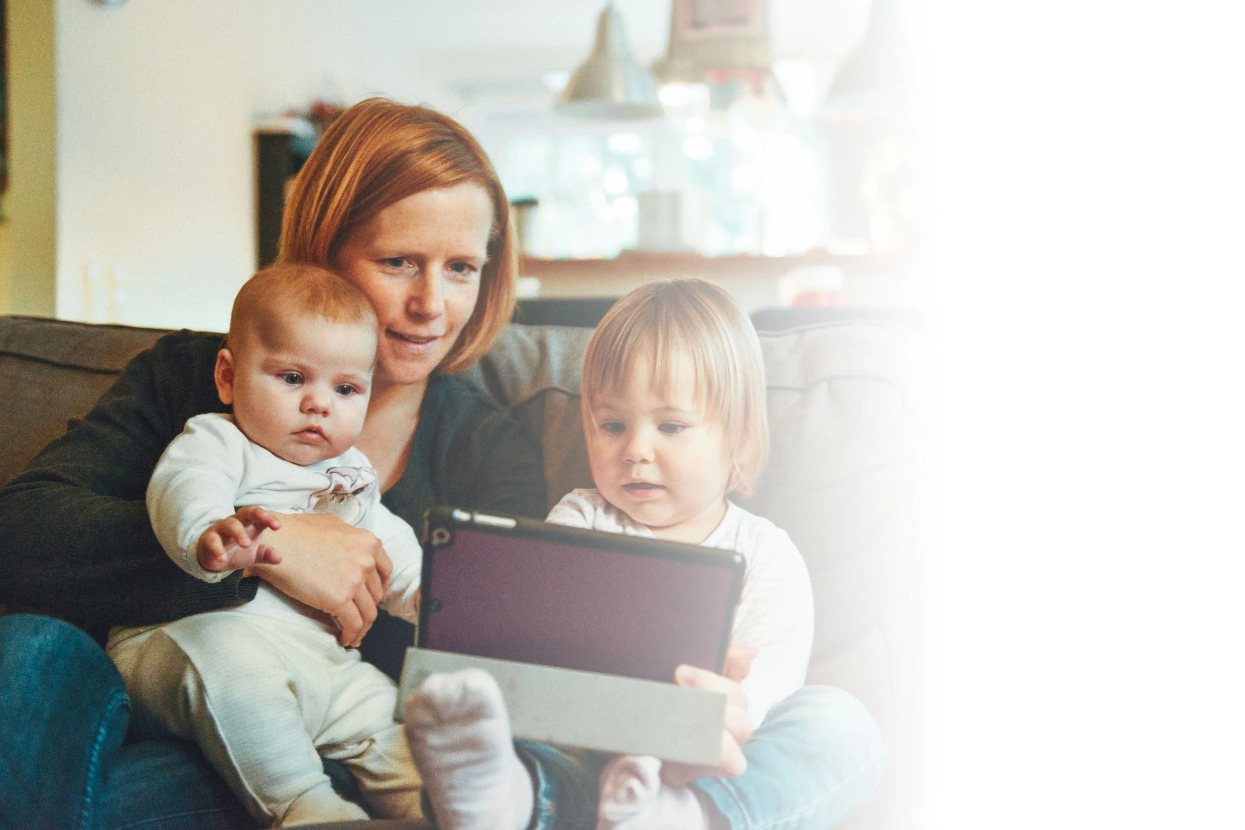 A woman with red hair sitting on a couch with two children, all looking at a tablet device.