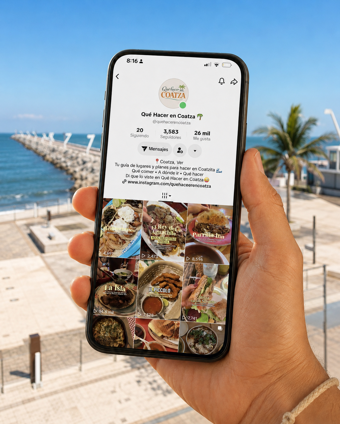 Persona sosteniendo un teléfono móvil mostrando una cuenta de Instagram de una página llamada 'Qué Hacer en Coatz' con fotos de comida y vistas al mar, con fondo de una playa y un muelle.