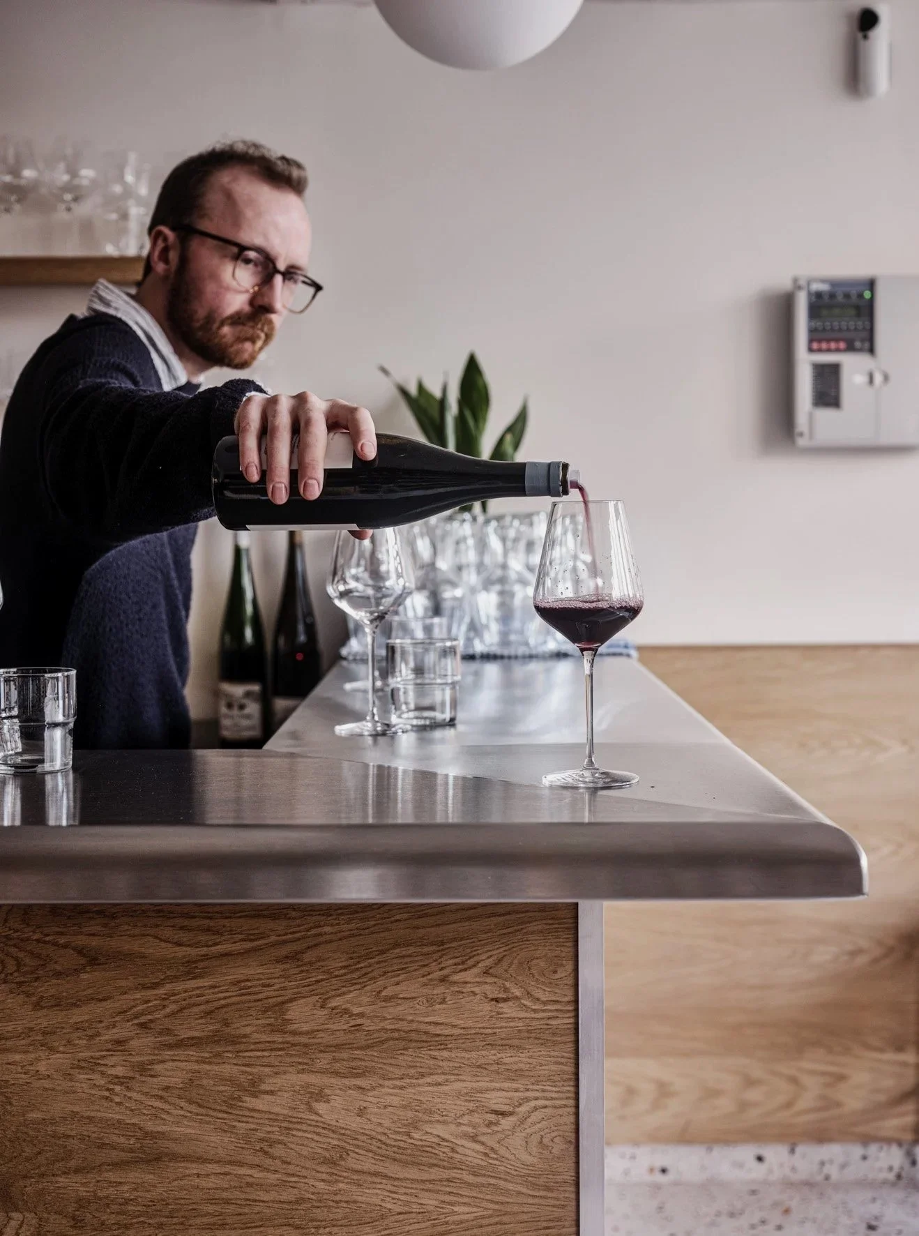 A man with glasses and a beard pouring red wine into a glass at a modern bar or kitchen counter. There are additional glasses and bottles in the background, and the wall features a plant and a control panel.