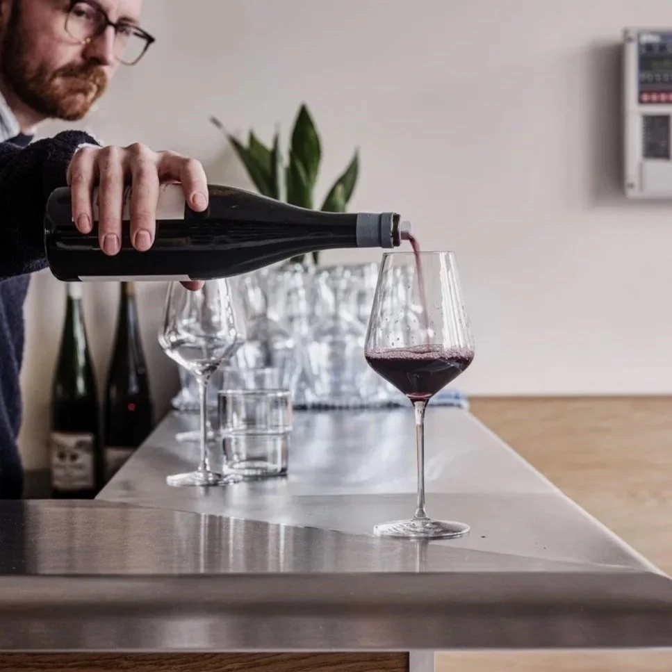 A man pouring red wine into a wine glass on a bar or counter, with empty glasses and bottles in the background.