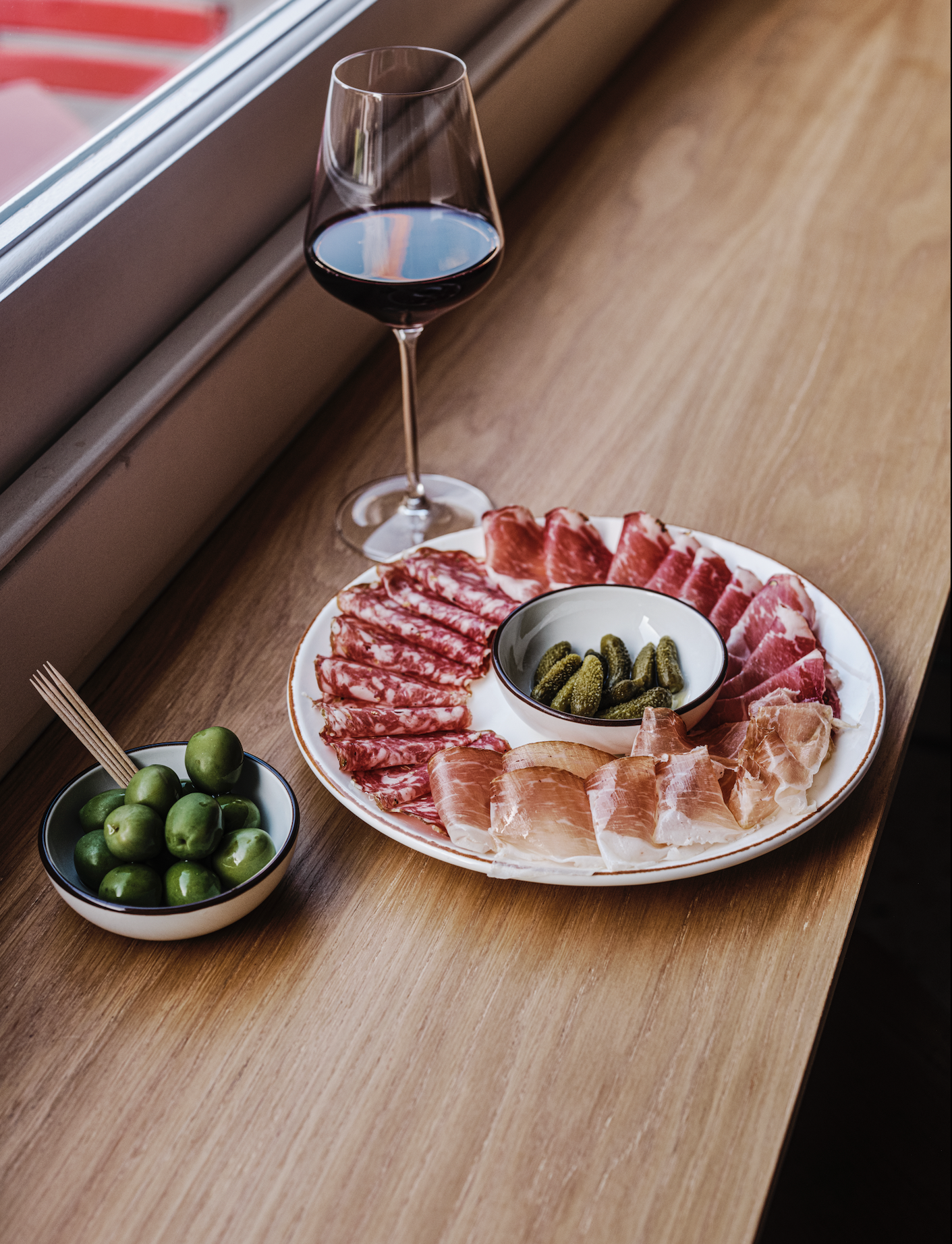 A wine glass filled with red wine, a plate of assorted cured meats including salami, prosciutto, and ham, a small bowl of green pickles, and a bowl of green olives on a wooden surface near a window.