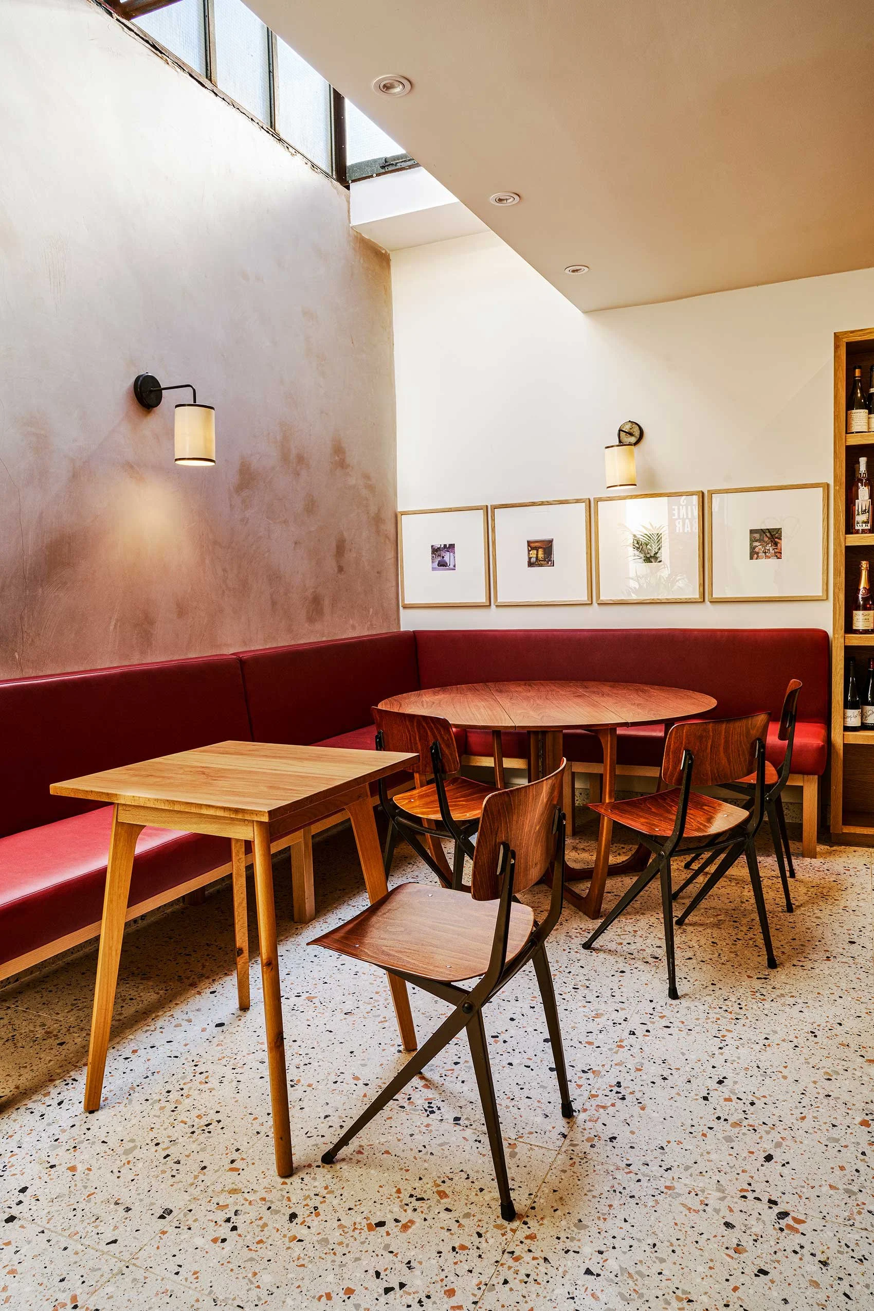 Interior of a modern restaurant with wooden tables, black and wooden chairs, a red banquette, framed photographs on the wall, and terrazzo flooring.