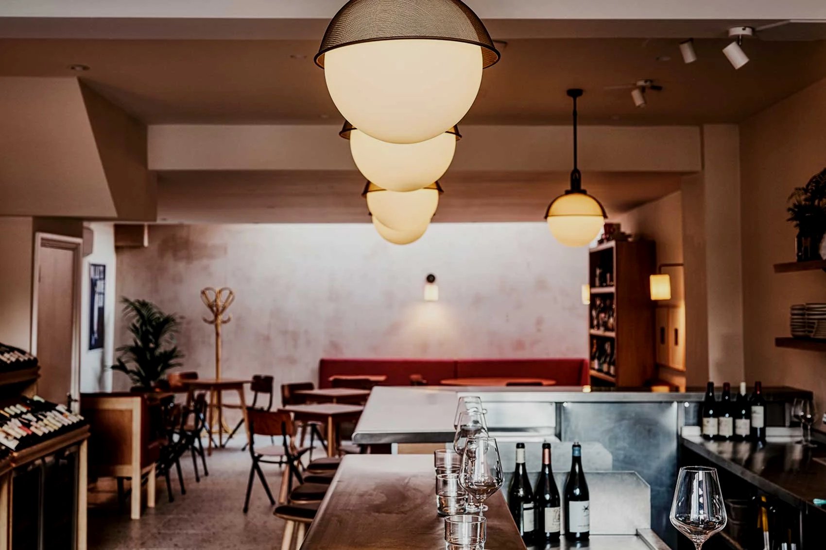 Interior of a cozy restaurant or cafe with warm lighting, wooden furniture, wine bottles on display, and decorative houseplants.
