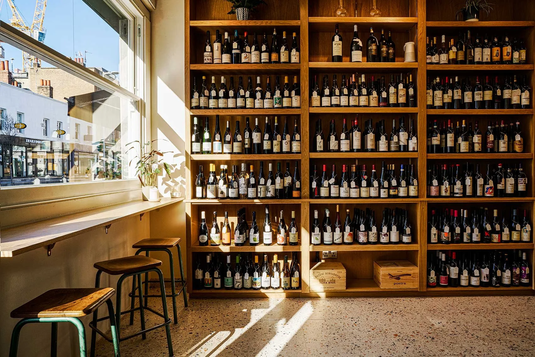A wall of wine bottles on wooden shelves in a cozy shop, with sunlight streaming through a nearby window, and a small counter with bar stools outside.