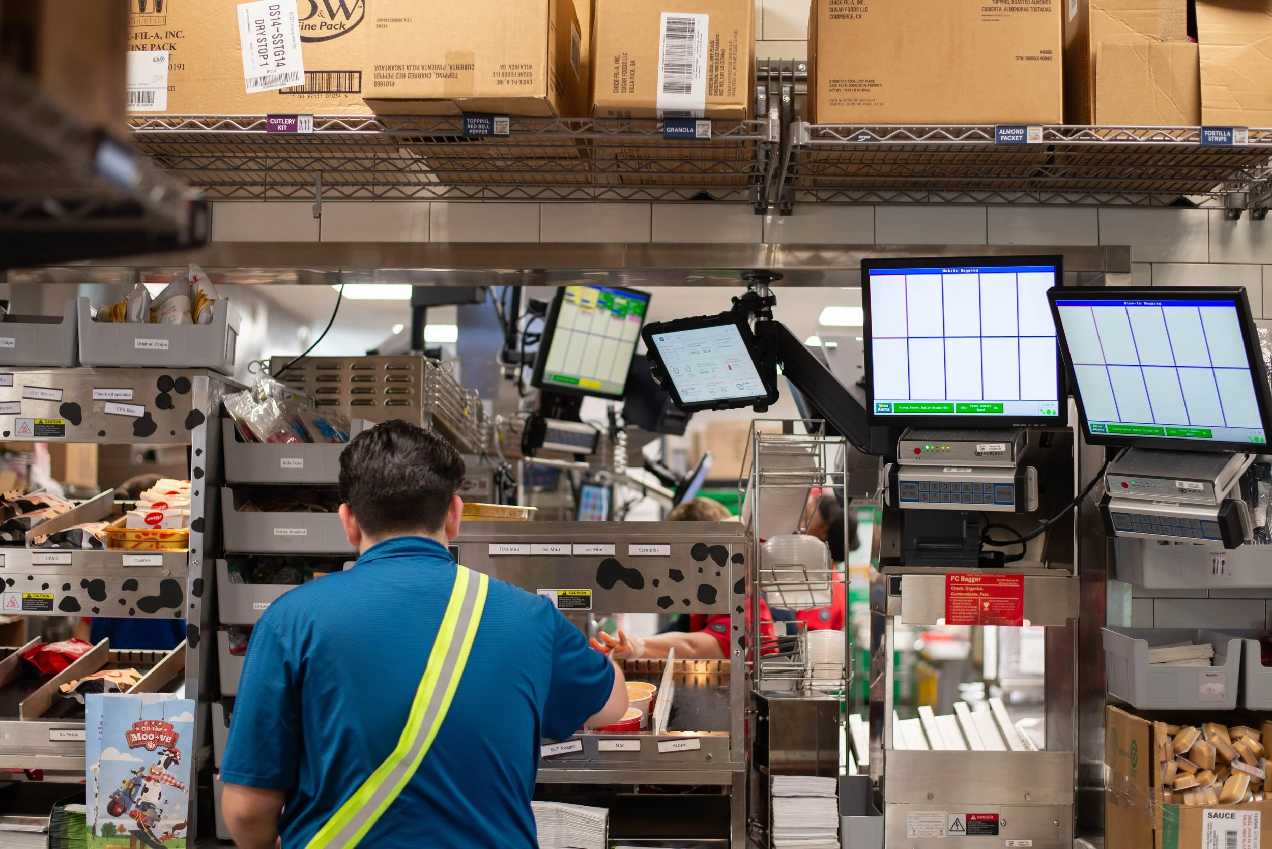 A worker in a blue uniform with yellow safety lines is working at a checkout counter in a grocery store. The background includes shelves with cardboard boxes, electronic checkout monitors, and various store items.