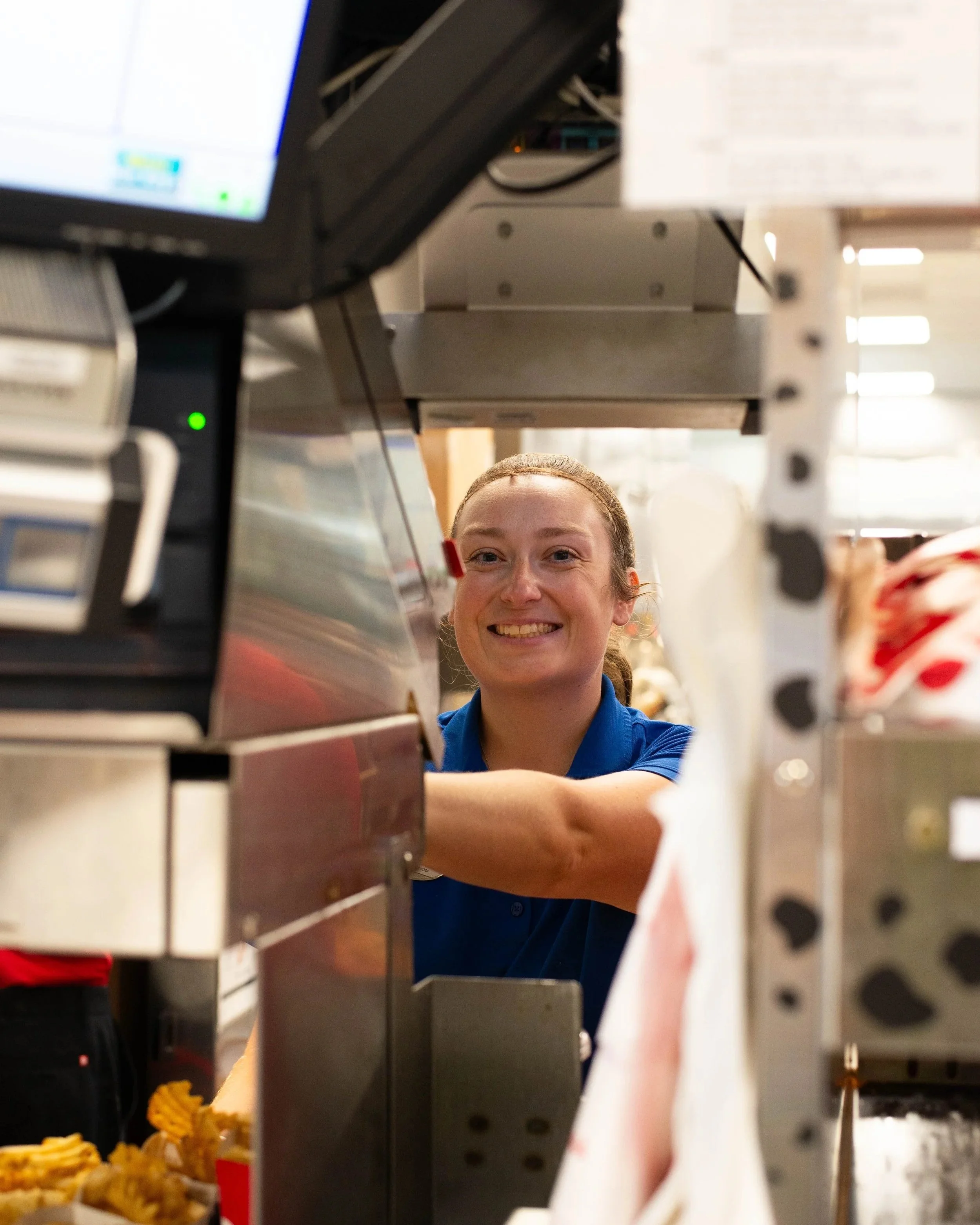 A woman in a blue uniform smiling behind a food service counter, with various kitchen equipment and food items visible in the background.