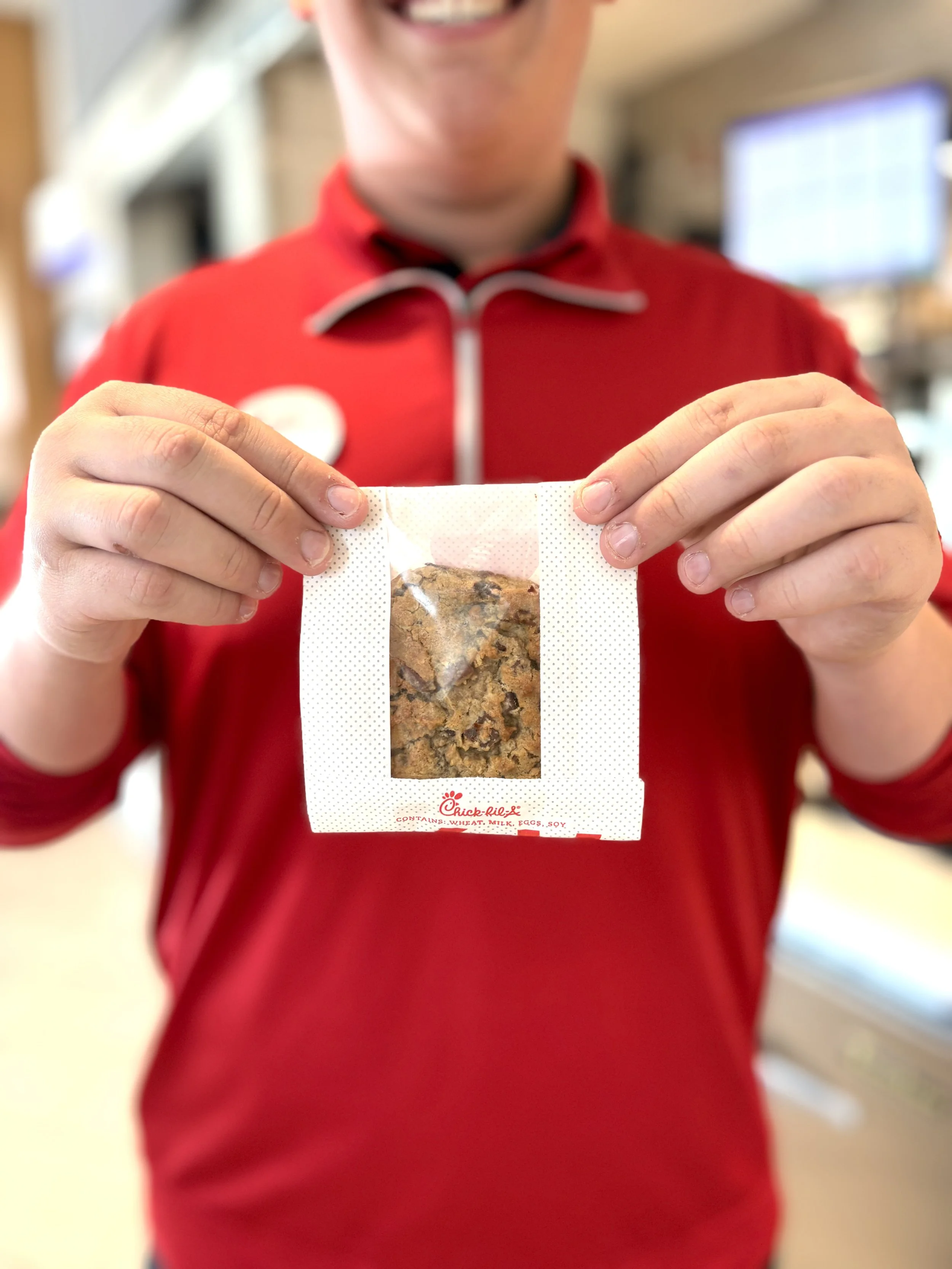 A person in a red shirt holding a Chick-fil-A cookie in front of the camera, with a blurred background.